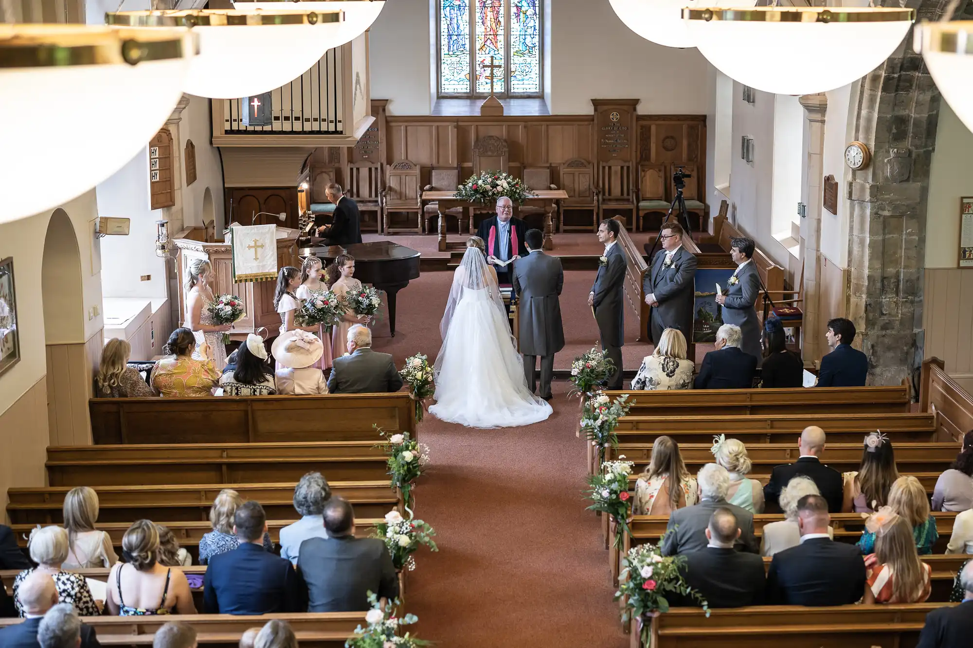 A bride and groom stand at the altar in a church, facing the officiant during a wedding ceremony. Guests are seated in pews, and bridal party members stand nearby.