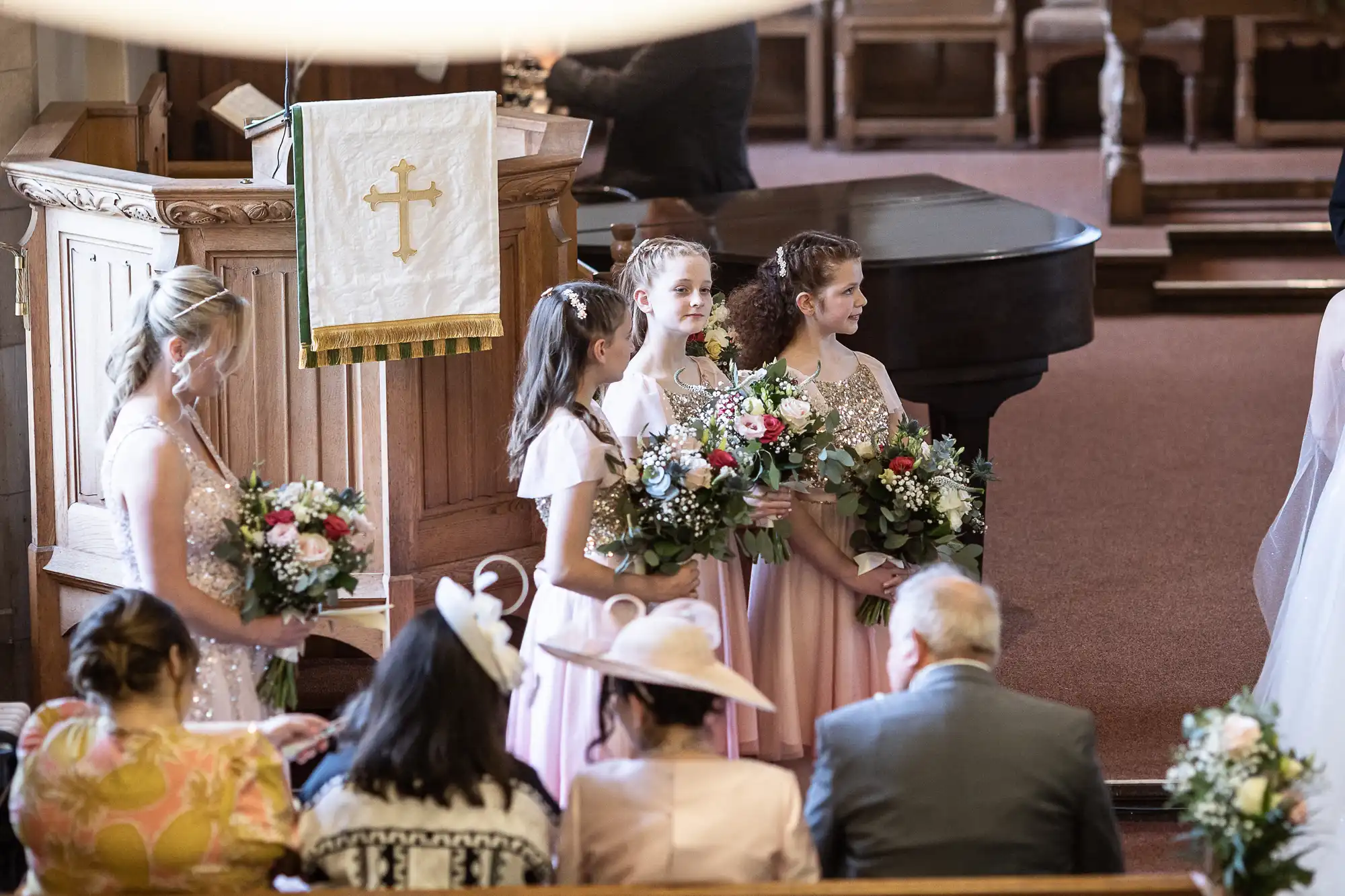 Four bridesmaids in light pink dresses and carrying flower bouquets stand at the front of a church. Guests are seated and an altar with a cross banner is in the background.