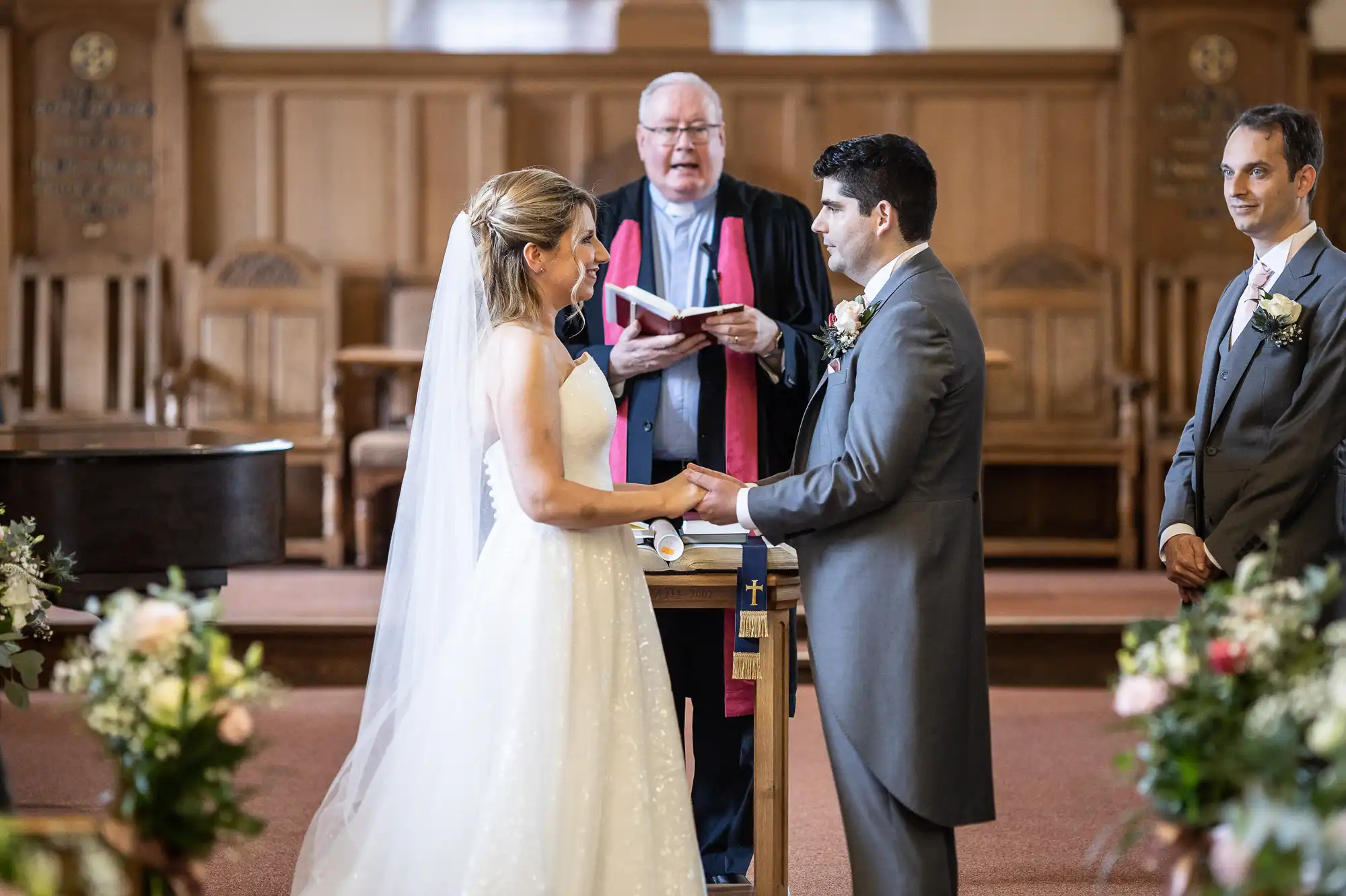 A bride and groom stand facing each other, holding hands, during a wedding ceremony officiated by a clergyman while a groomsman looks on.