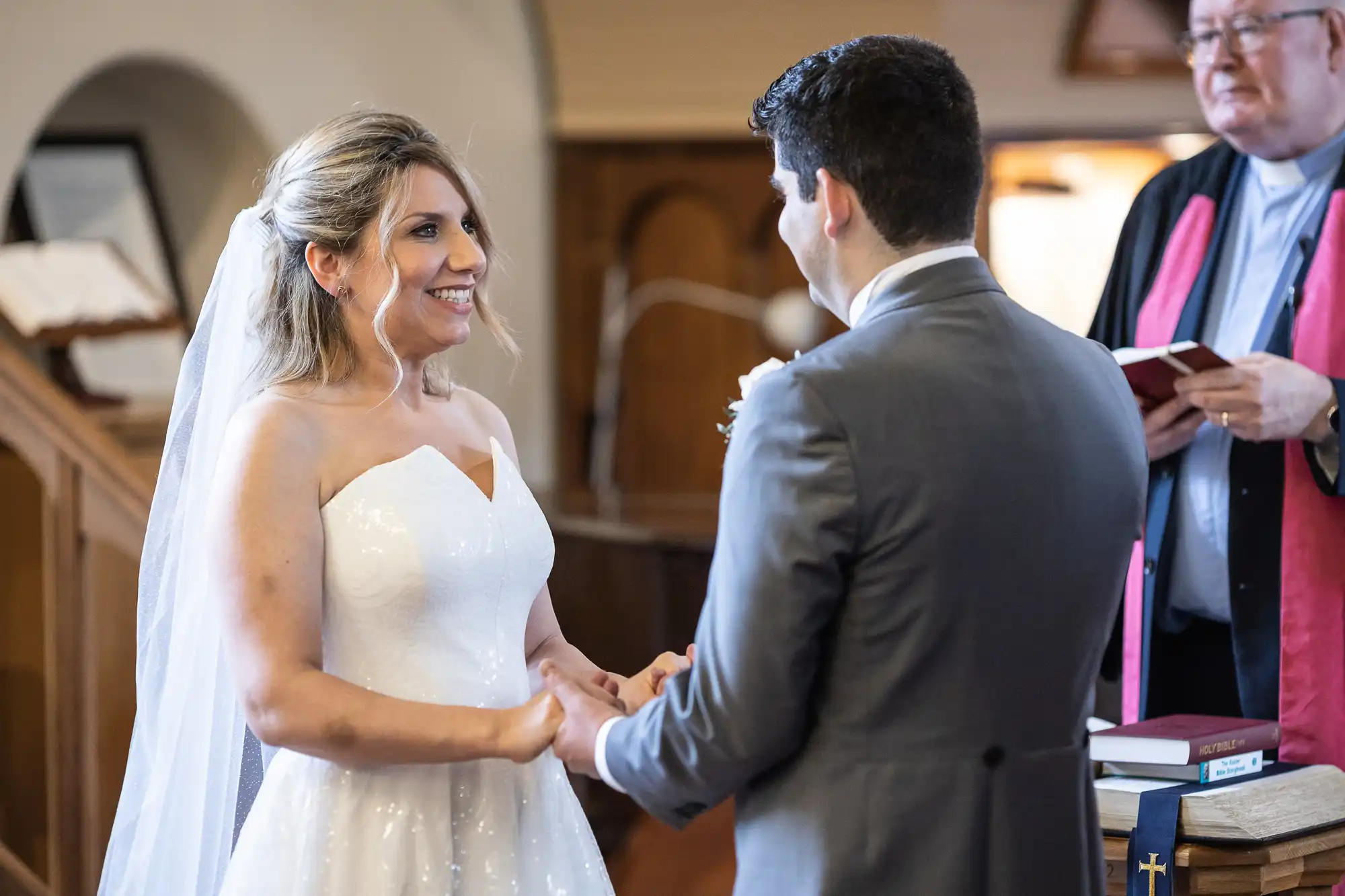 A bride and groom hold hands and smile at each other during a wedding ceremony, with an officiant standing nearby holding a book.