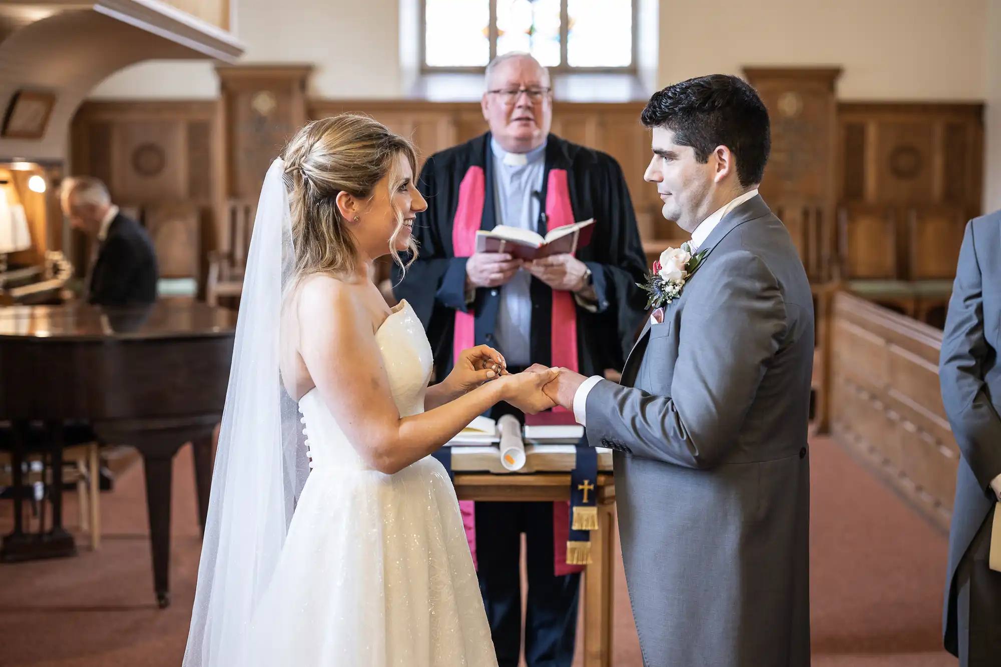 A bride and groom exchange rings during their wedding ceremony in a church, with an officiant standing behind them holding a book. The bride wears a white dress and veil; the groom wears a gray suit.