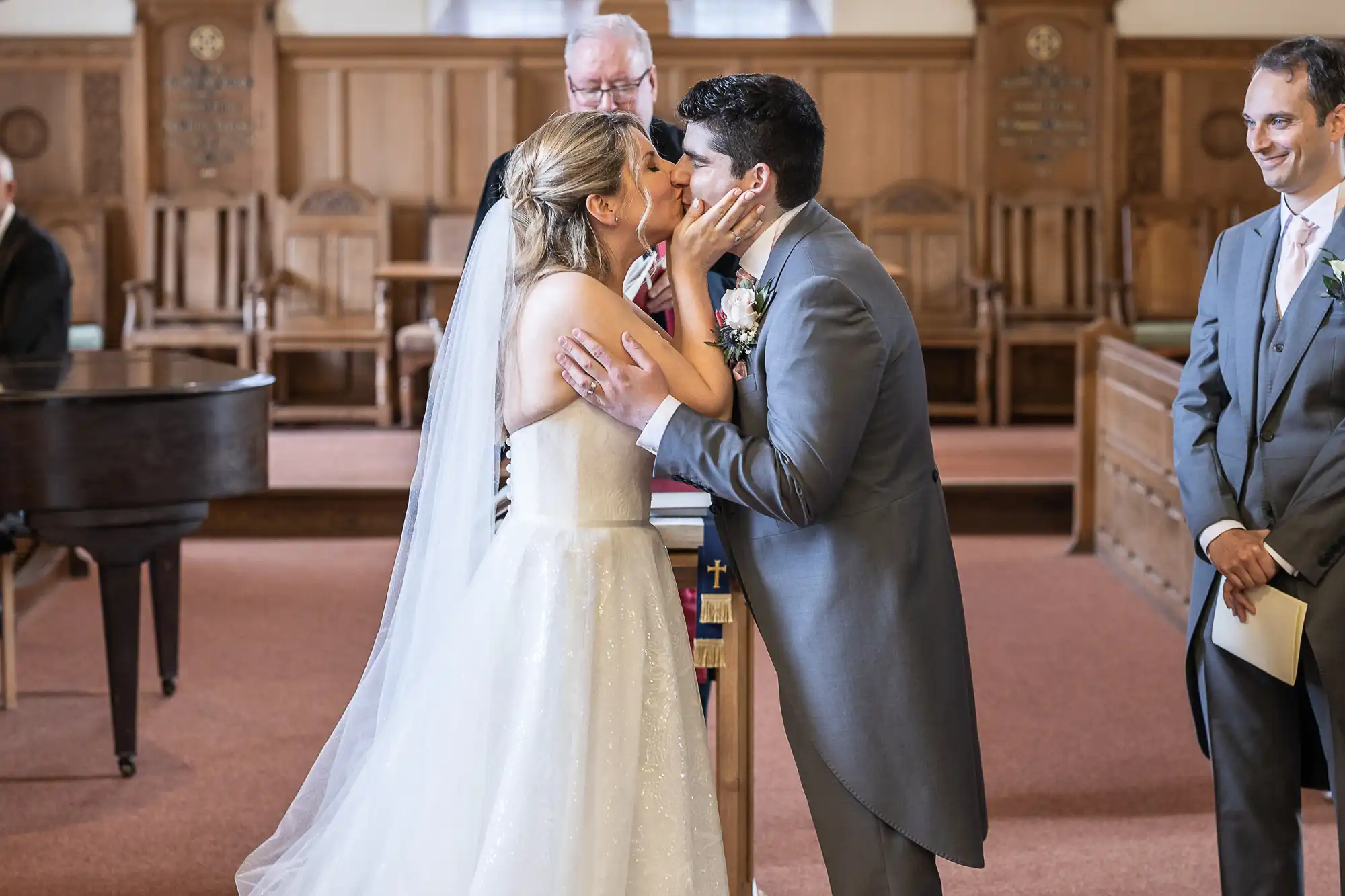 A bride and groom share a kiss at their wedding ceremony in a church, with an officiant and a groomsman standing nearby. The bride wears a white gown and veil, and the groom is in a gray suit.