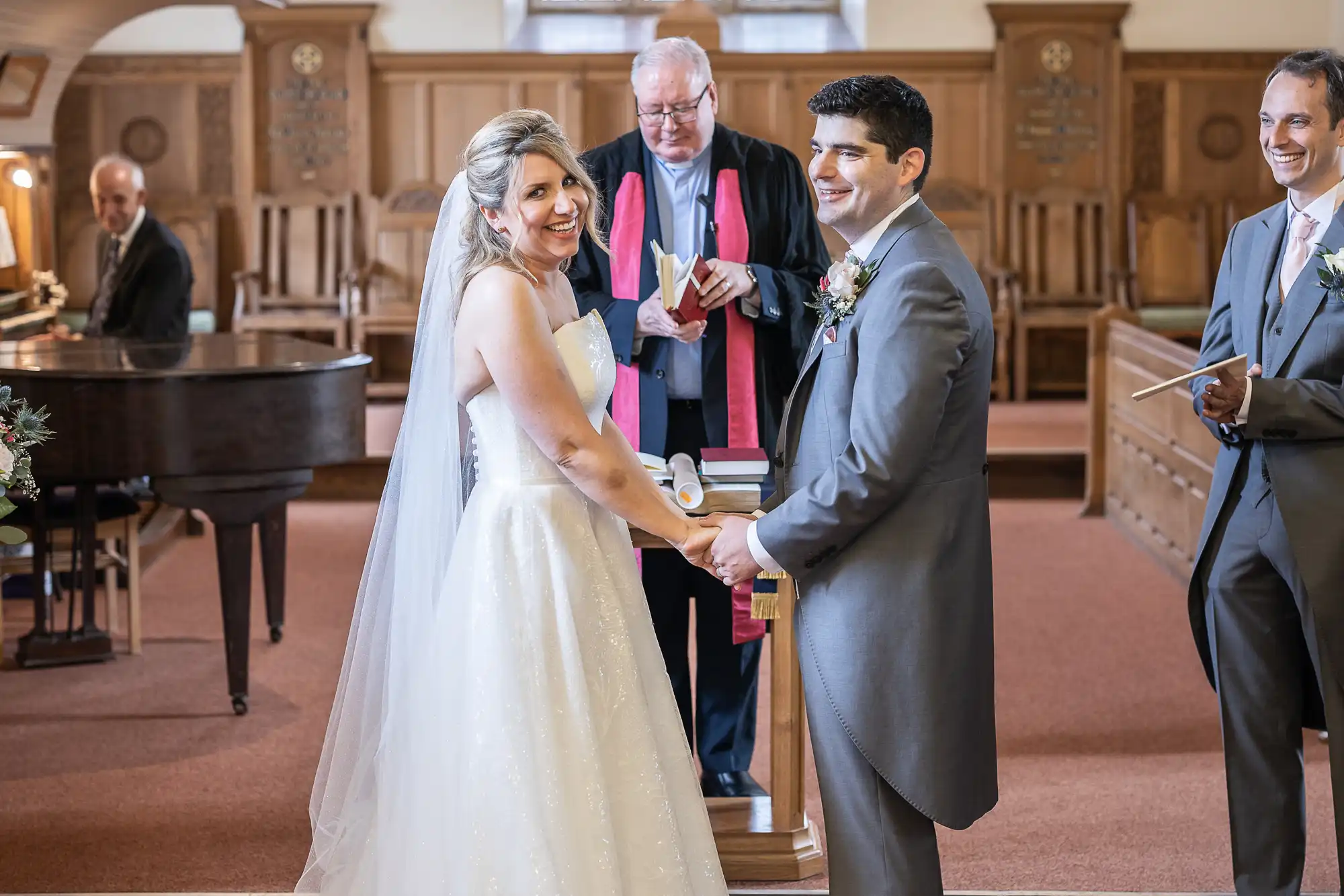 A bride and groom smile and hold hands at the altar during their wedding ceremony in a church, with the officiant and groomsmen standing behind them.