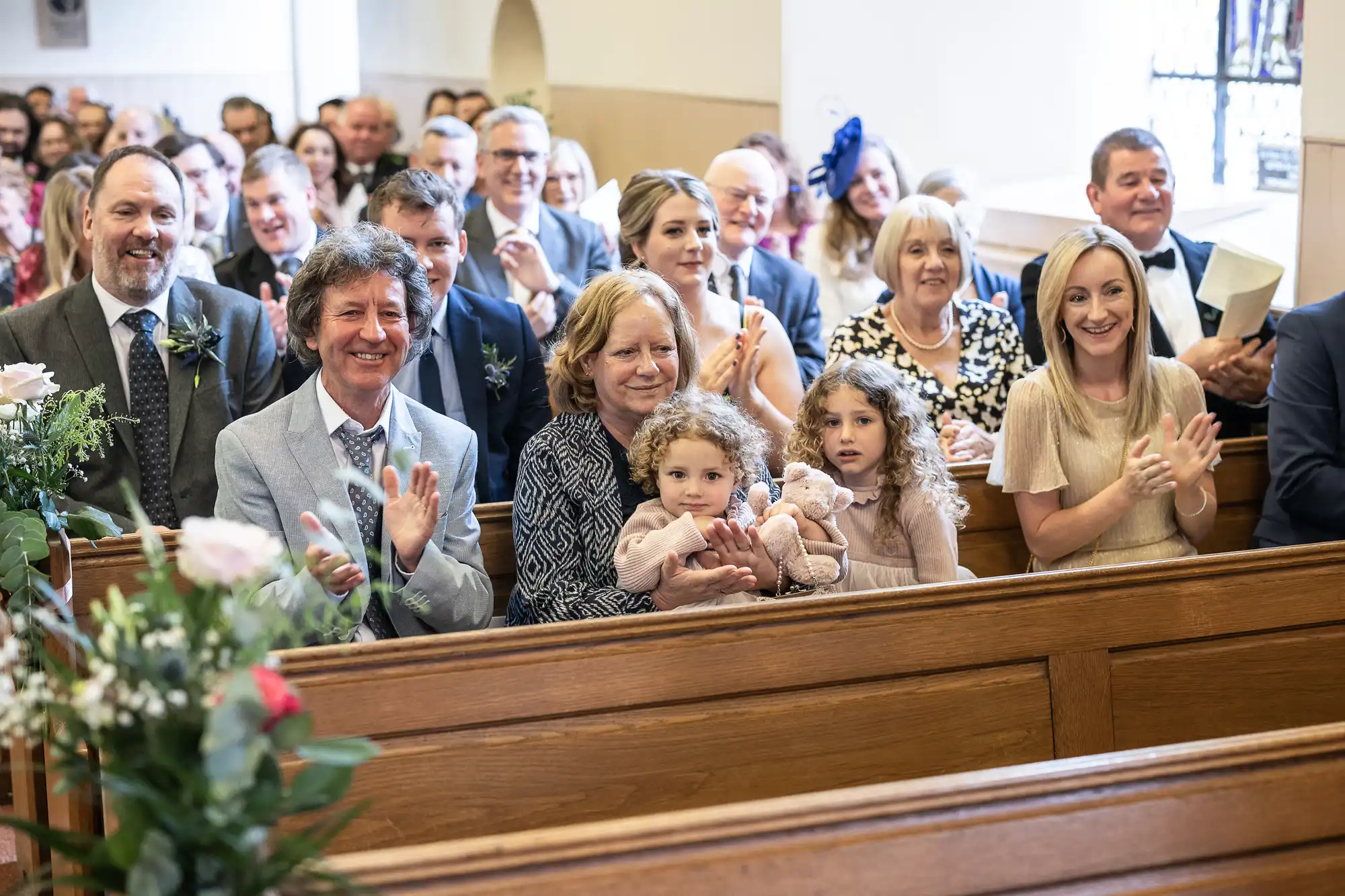 A group of people seated in pews at a church event, smiling and clapping. Two young children are among the attendees, one of whom is holding a soft toy.