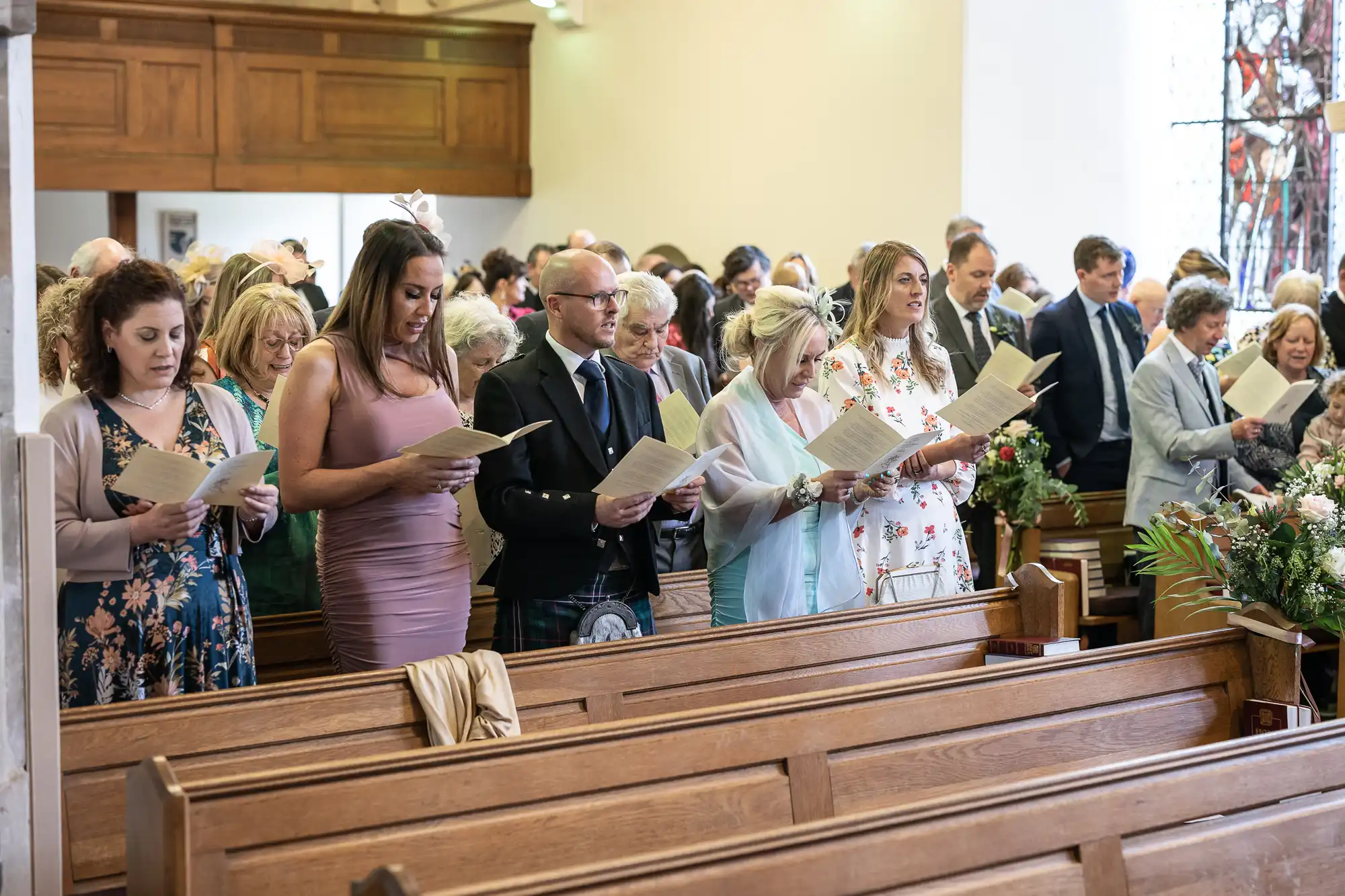 A group of people, dressed formally, stand in a church holding hymn sheets. Some are wearing floral dresses, suits, and other formal attire, while attending a gathering or ceremony inside the church.