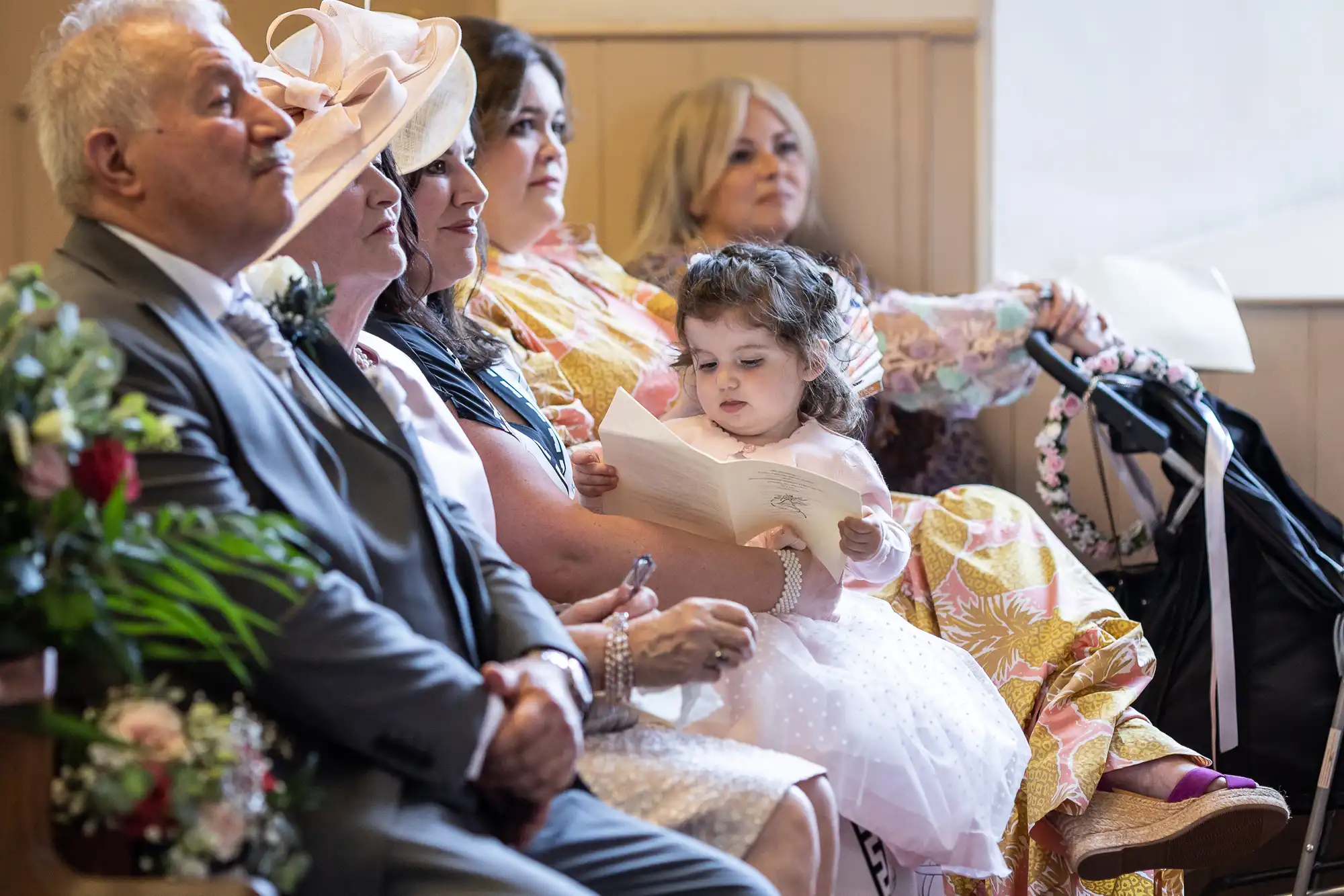 Several adults and a young girl sit in a row during a formal event. The girl is holding and looking at a booklet. A stroller is visible on the right.