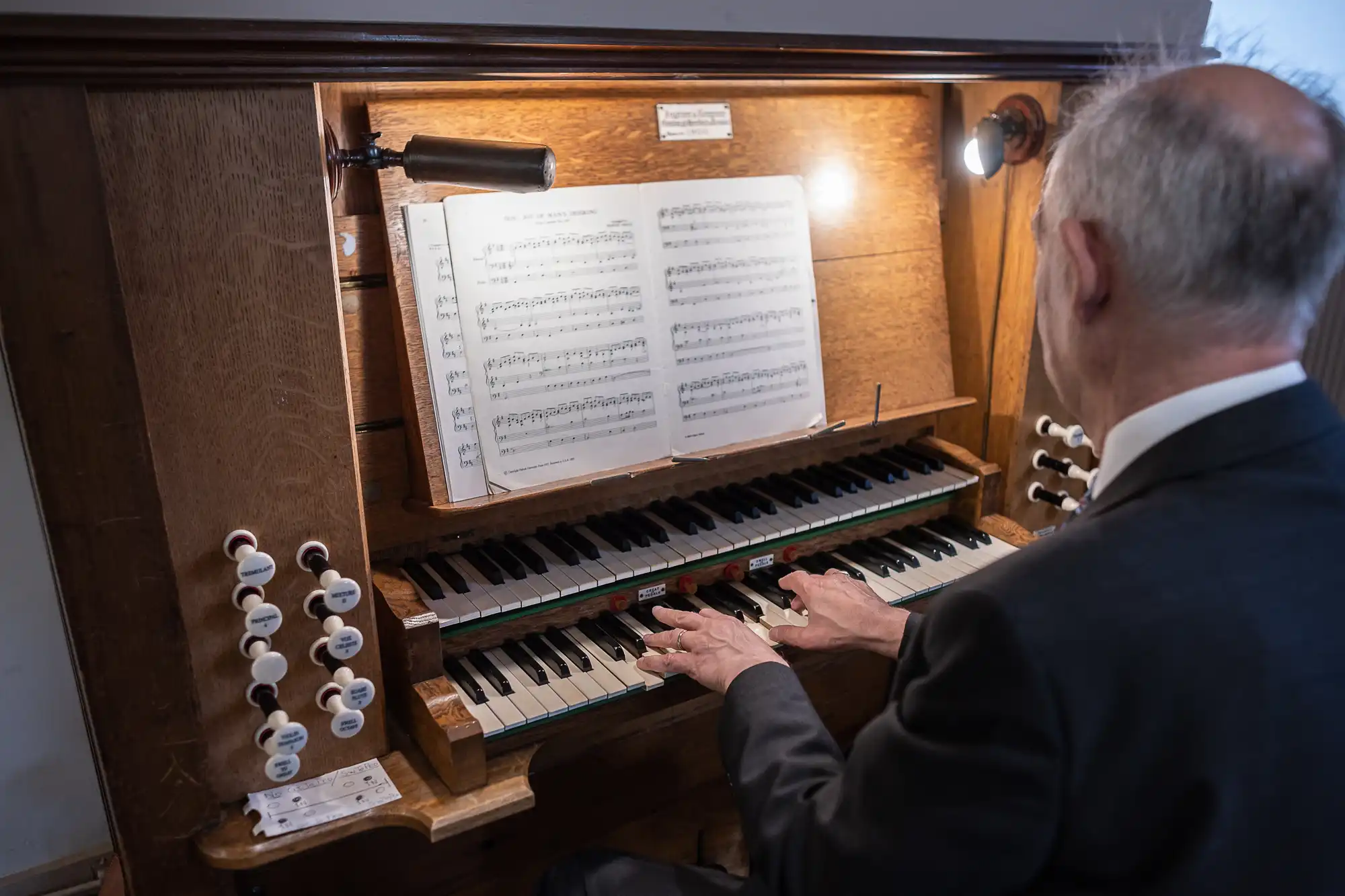 A man in a suit plays a pipe organ, reading sheet music. His fingers are on the keyboards, and multiple stops are visible beside the keyboards. Two lights illuminate the sheet music.