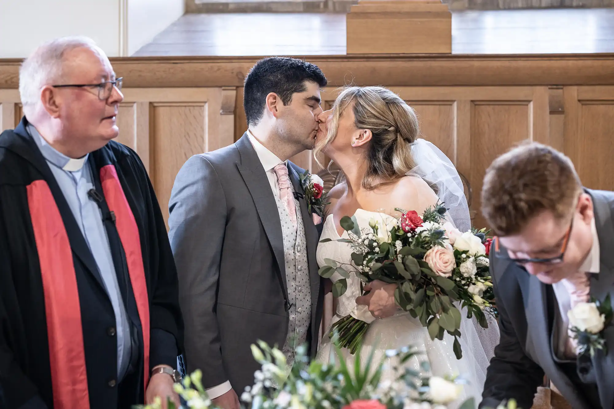 A couple in wedding attire kisses at the altar. A man in a suit stands nearby, and another individual in a suit signs a document. There are flowers in the foreground.