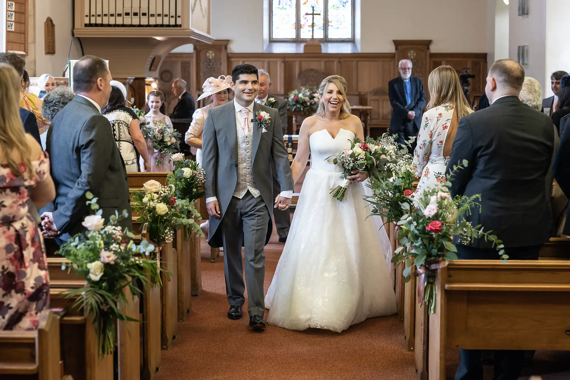 A bride and groom walk down the aisle smiling, hand-in-hand, in a church decorated with flowers and greenery, surrounded by standing guests.