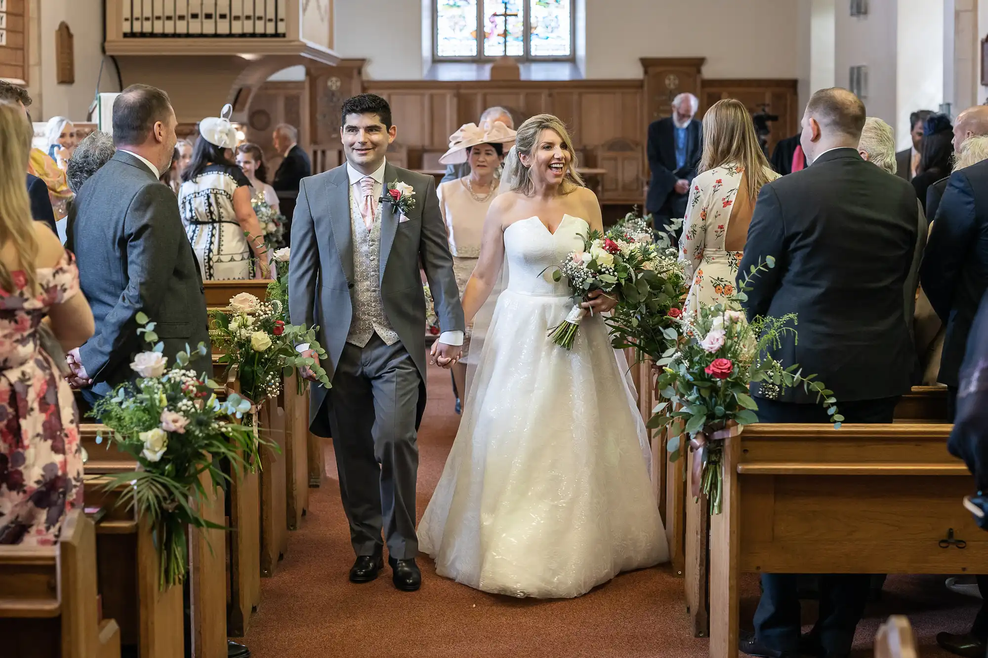 A bride and groom walk down the aisle, smiling and holding hands, surrounded by guests in a church decorated with flowers.