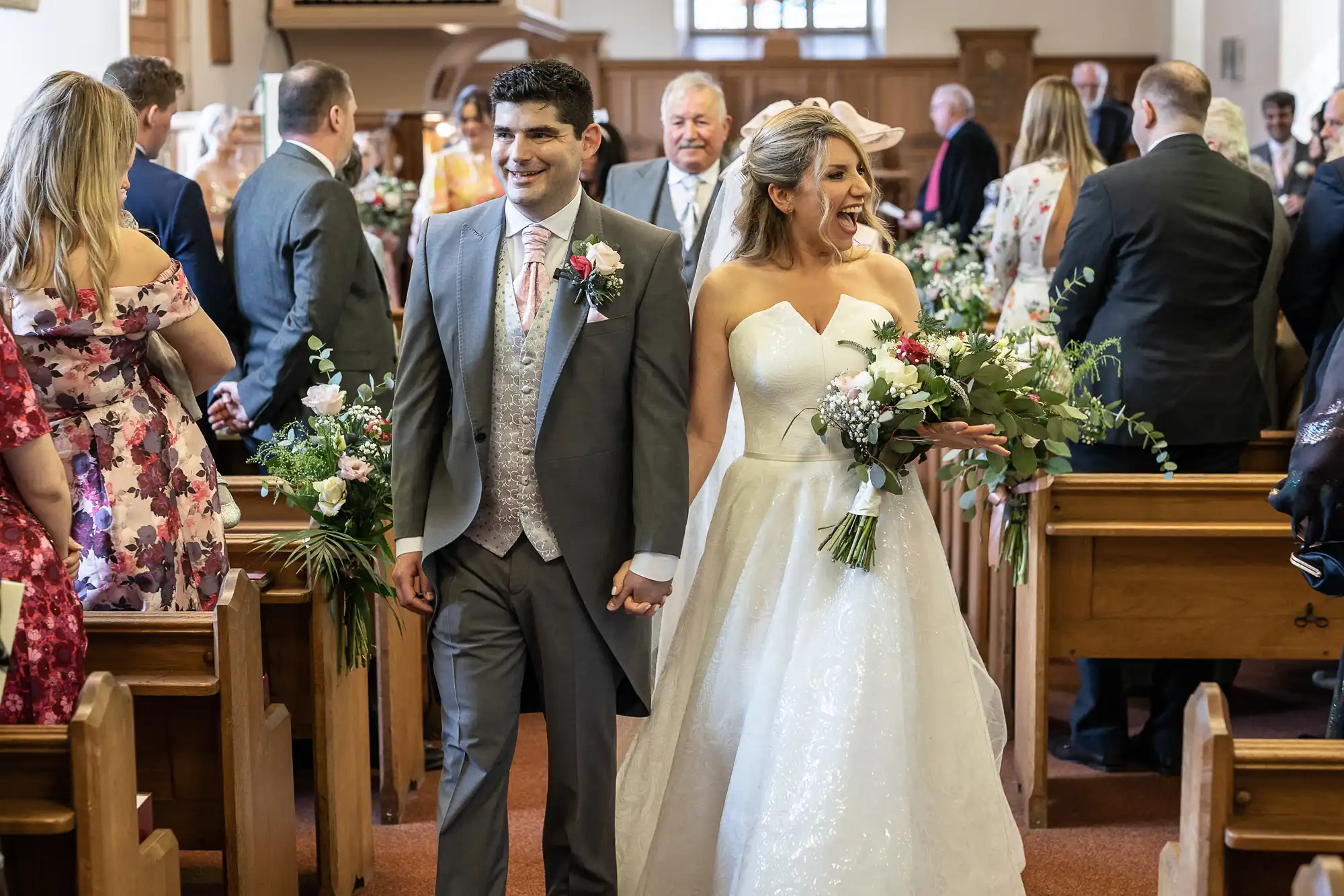 A bride and groom are walking down the aisle, smiling and holding hands, surrounded by guests inside a church. The bride holds a bouquet and wears a white dress, while the groom is in a grey suit.