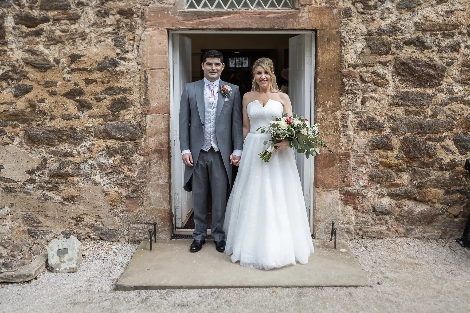 A bride and groom stand holding hands in a doorway, both smiling at the camera. The groom is dressed in a grey suit, and the bride is wearing a white wedding dress and holding a bouquet of flowers.