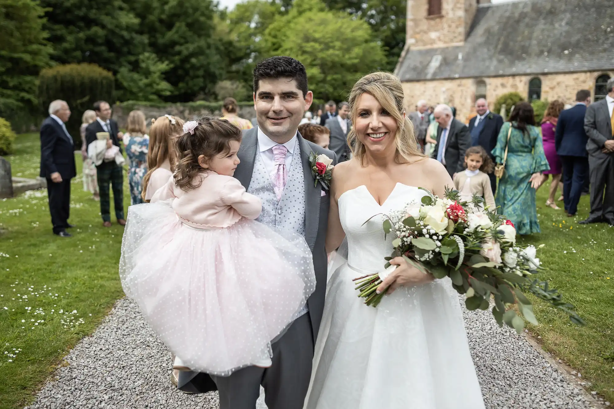 A bride and groom stand outdoors posing for a photo, the groom holding a young child. A group of people is gathered in the background near a stone building.