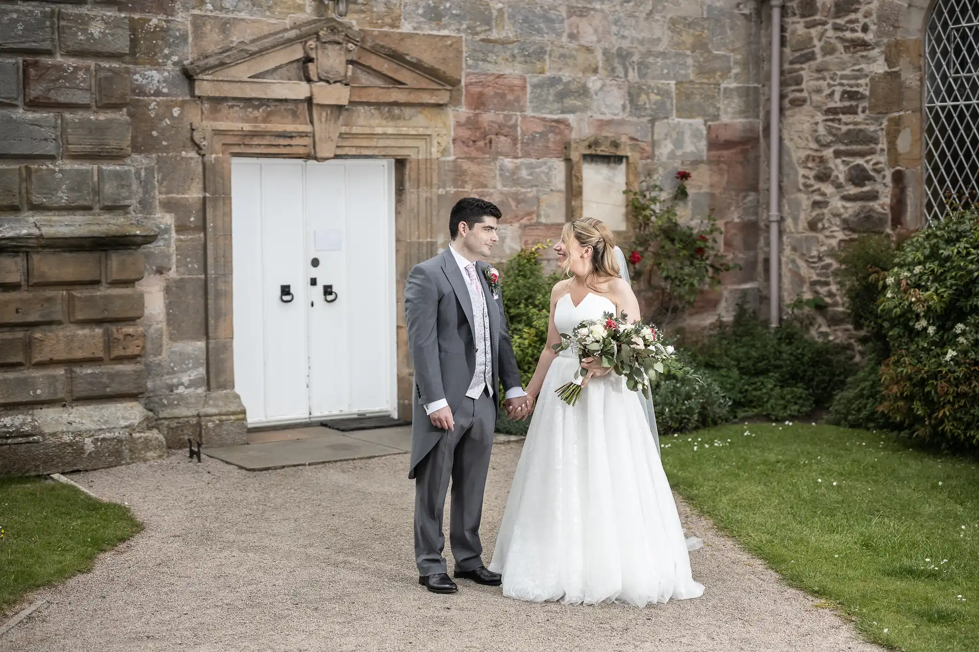 A bride in a white gown and a groom in a gray suit stand holding hands in front of a stone building with a white door and a small garden.