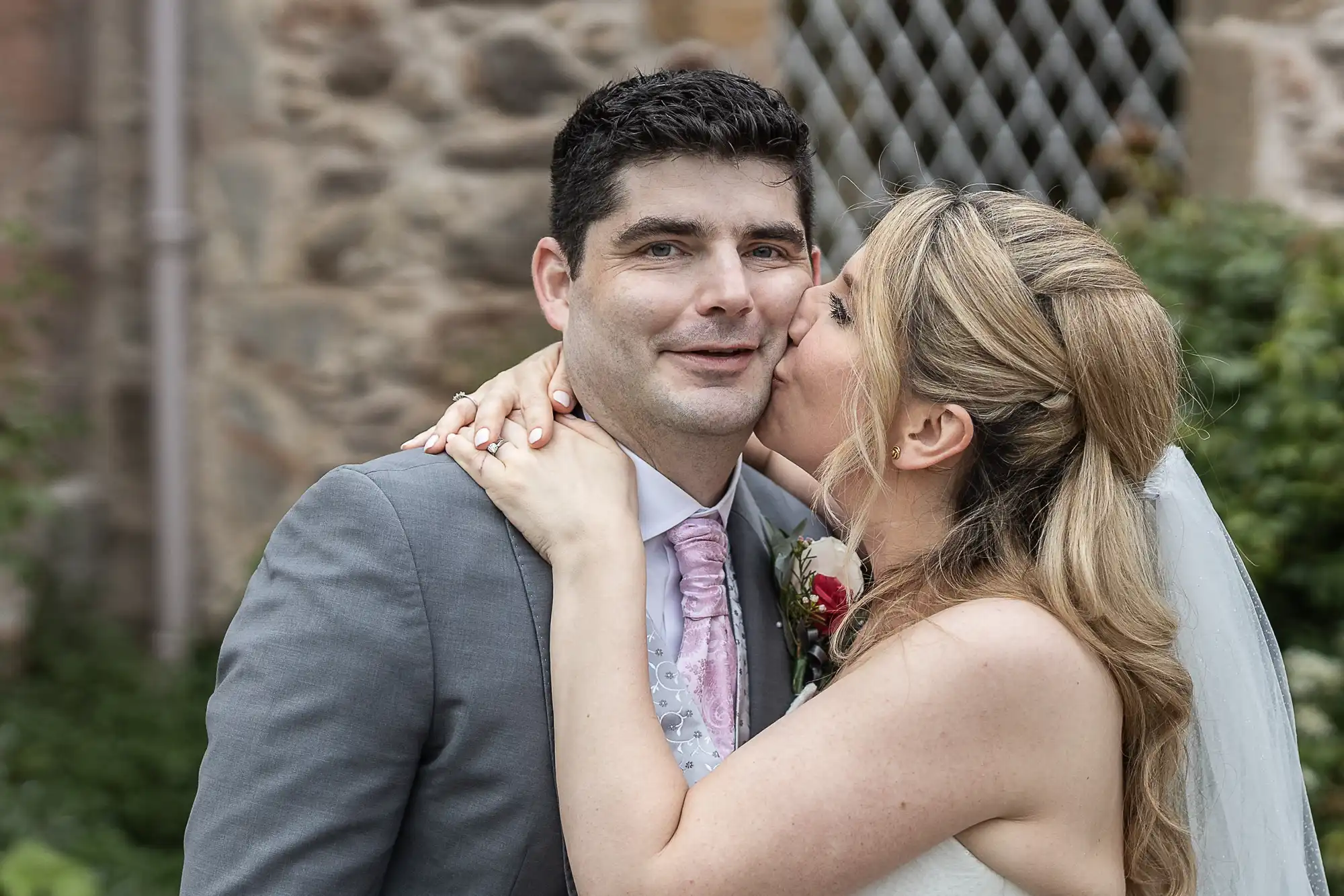 A man in a gray suit smiles as a woman in a white dress and veil kisses his cheek. They stand outdoors in front of a stone wall and lattice window.