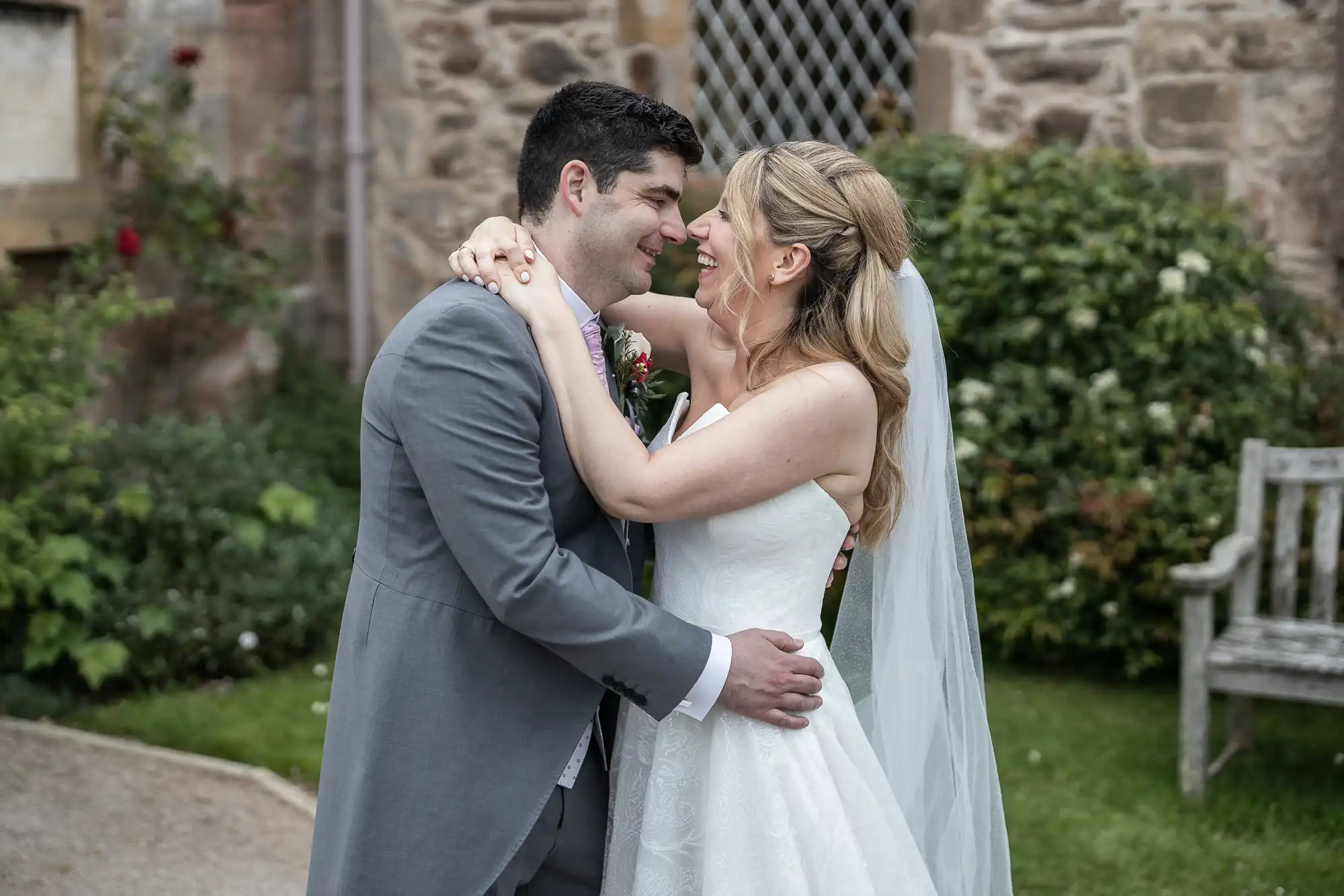 Marine Hotel wedding photos: A newlywed couple in wedding attire embraces outdoors near a stone building, smiling at each other. The groom wears a grey suit, and the bride wears a white dress with a veil.