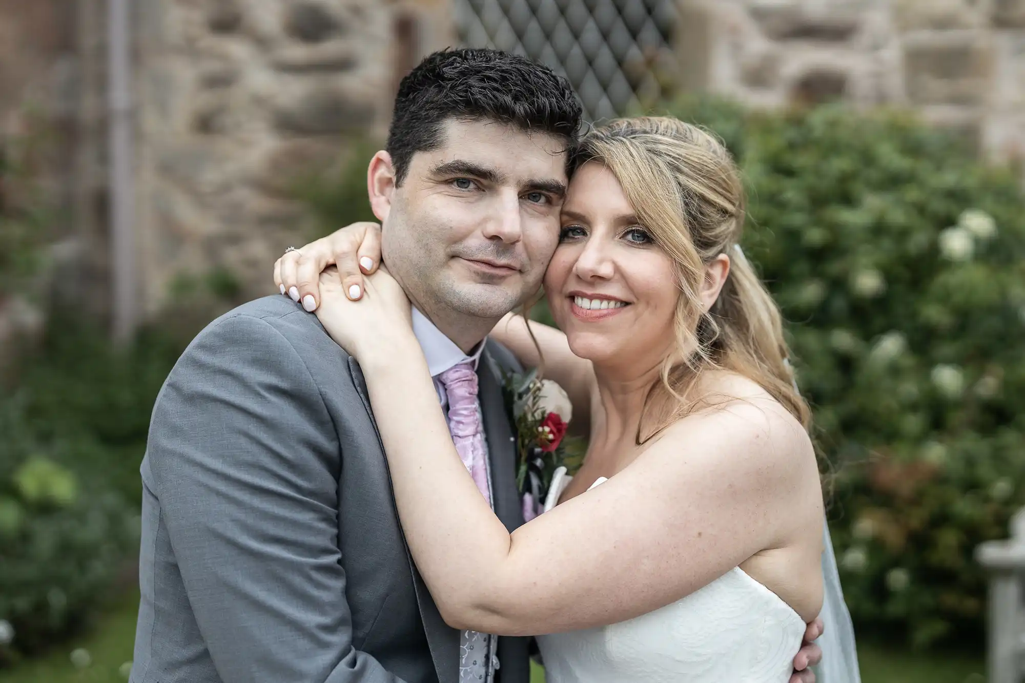 A couple stands close together outside, with the woman's arms around the man's neck. He wears a gray suit and she is in a white dress. They both smile at the camera.