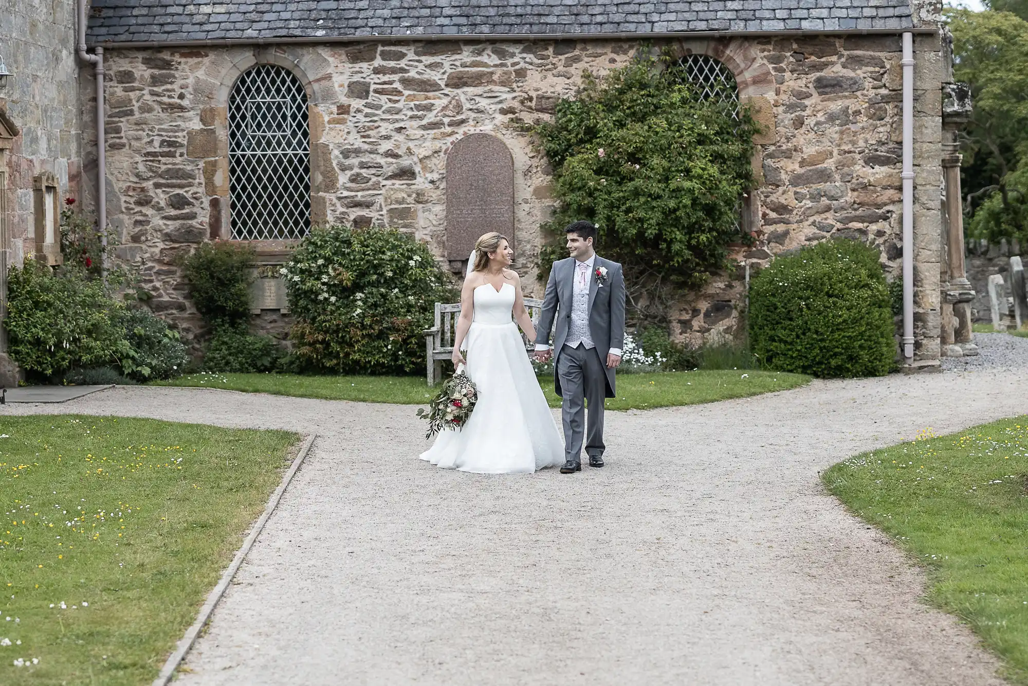 A bride and groom walk hand-in-hand outside a stone church on a gravel path, with greenery and lawn on either side.