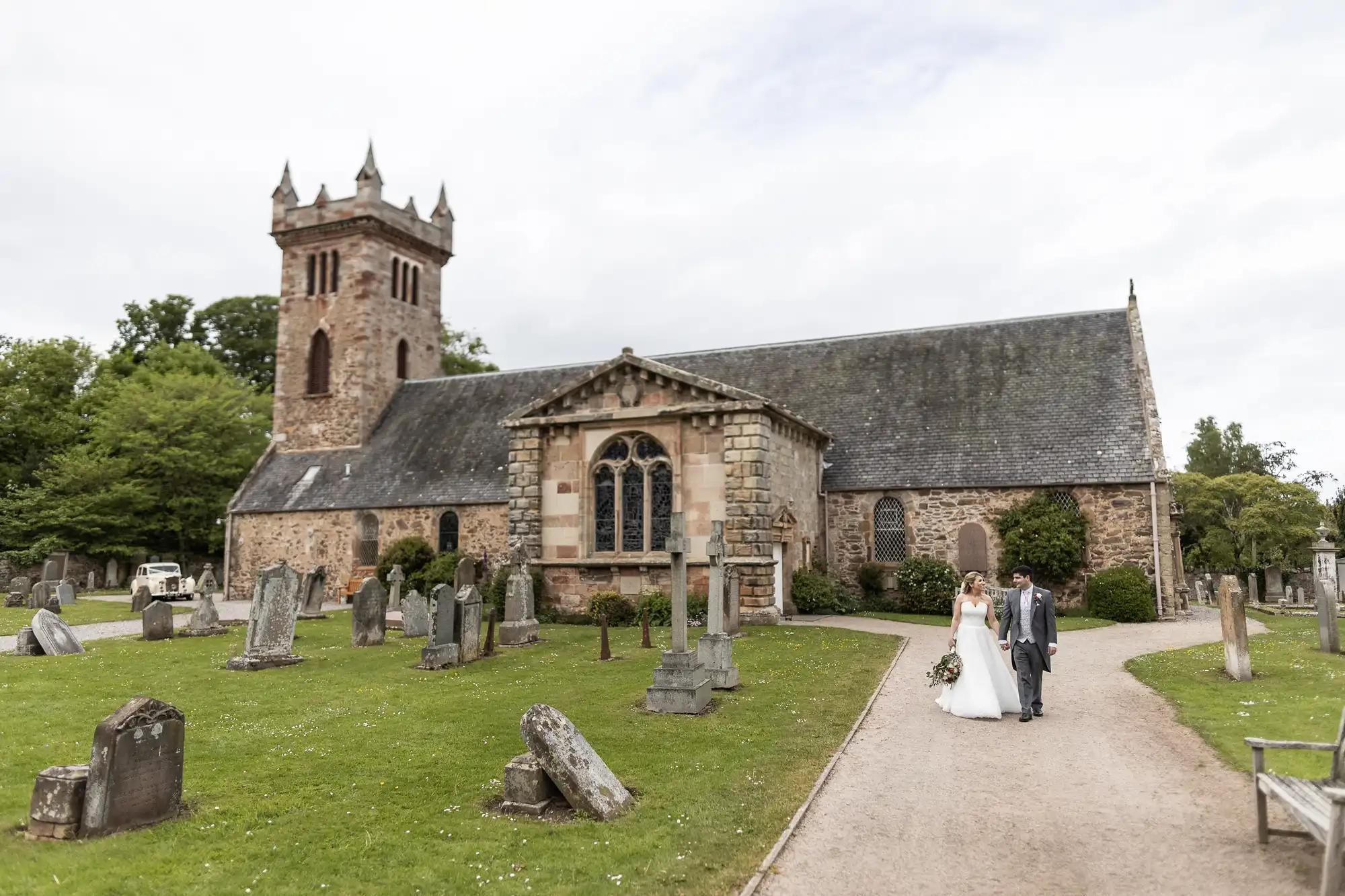 A bride and groom walk hand-in-hand on a gravel path in front of an old stone church with a cemetery surrounding it.