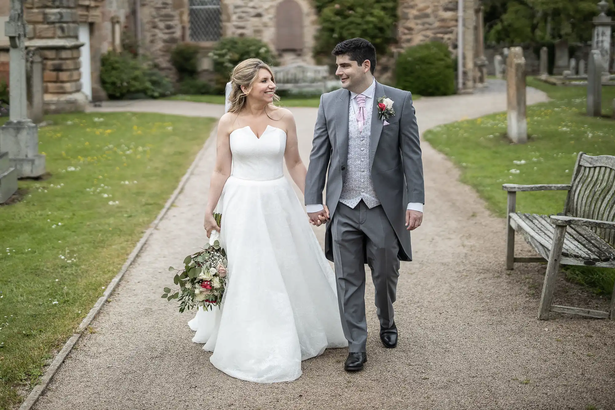 A couple in wedding attire walks hand in hand on a path through a garden with medieval buildings in the background. The bride holds a bouquet and wears a white gown, while the groom wears a grey suit.