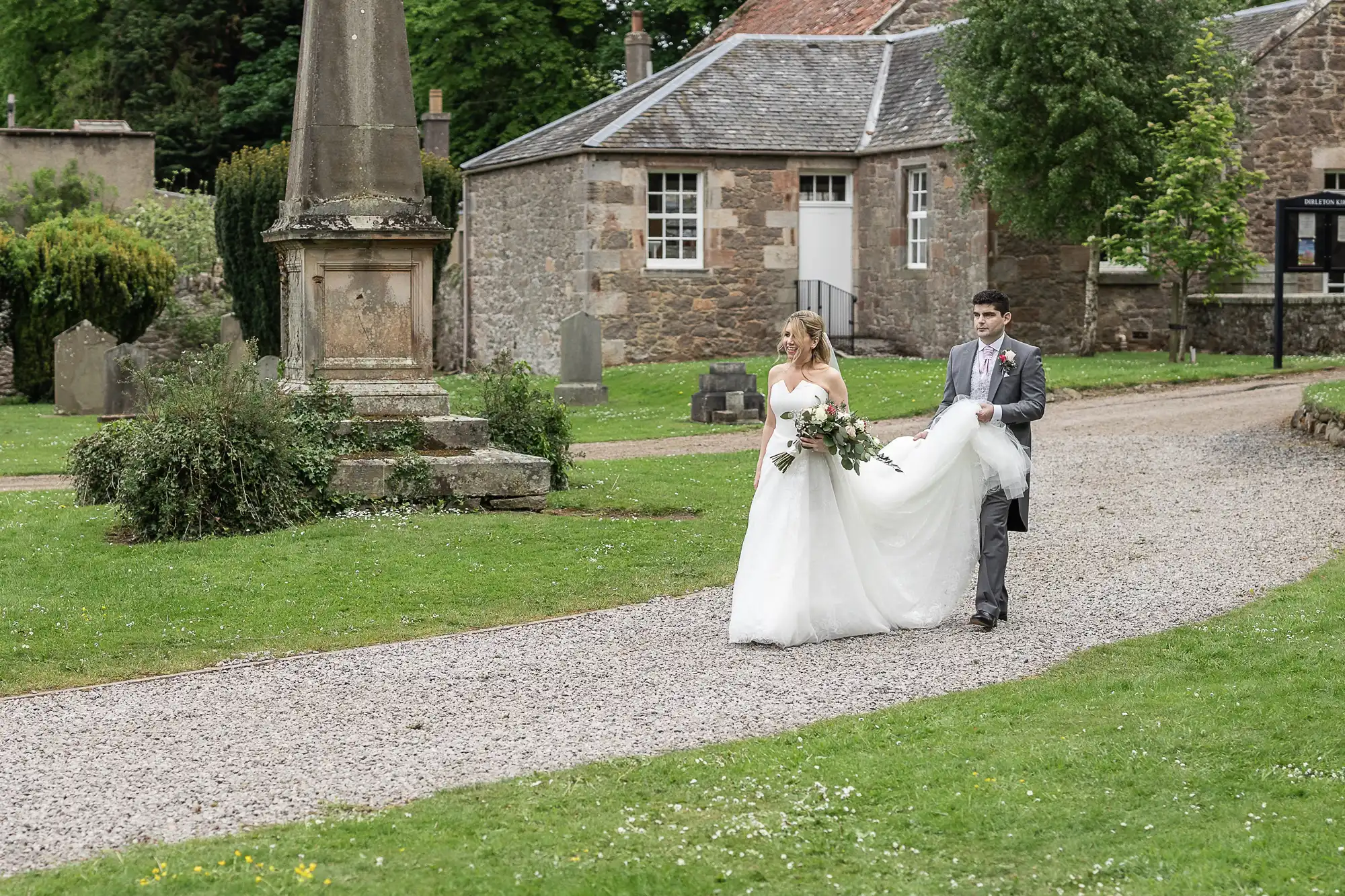 A bride and groom walk together on a gravel path through a garden with historic stone buildings in the background. The groom is holding the train of the bride's wedding dress.