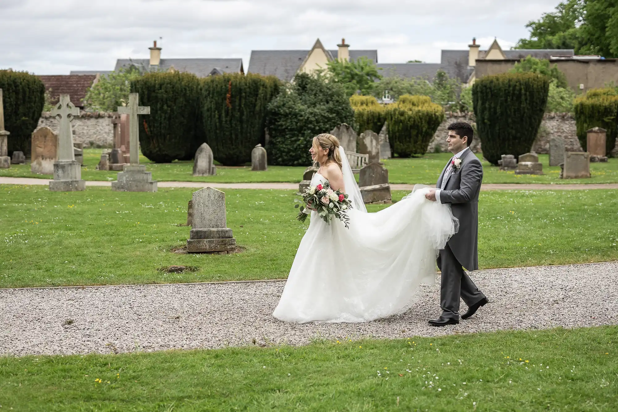 A bride and groom walk through a cemetery. The bride holds a bouquet and the groom lifts the bride's train. Gravestones and green grass are in the background.