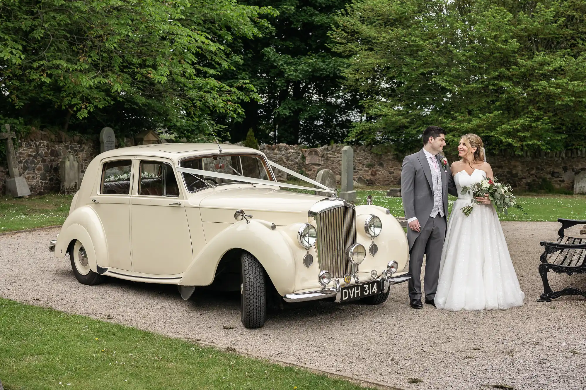 Bride and groom stand beside a vintage ivory car in a garden setting. The bride holds a bouquet and the groom wears a gray suit. Trees and stone structures are visible in the background.