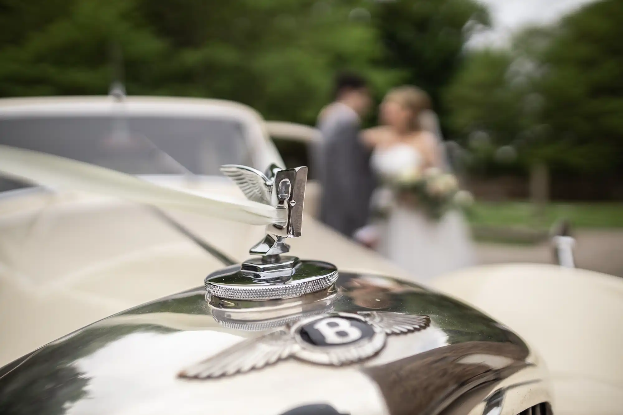 Close-up of a vintage Bentley hood ornament with a ribbon. In the blurred background, a bride and groom stand together outdoors.