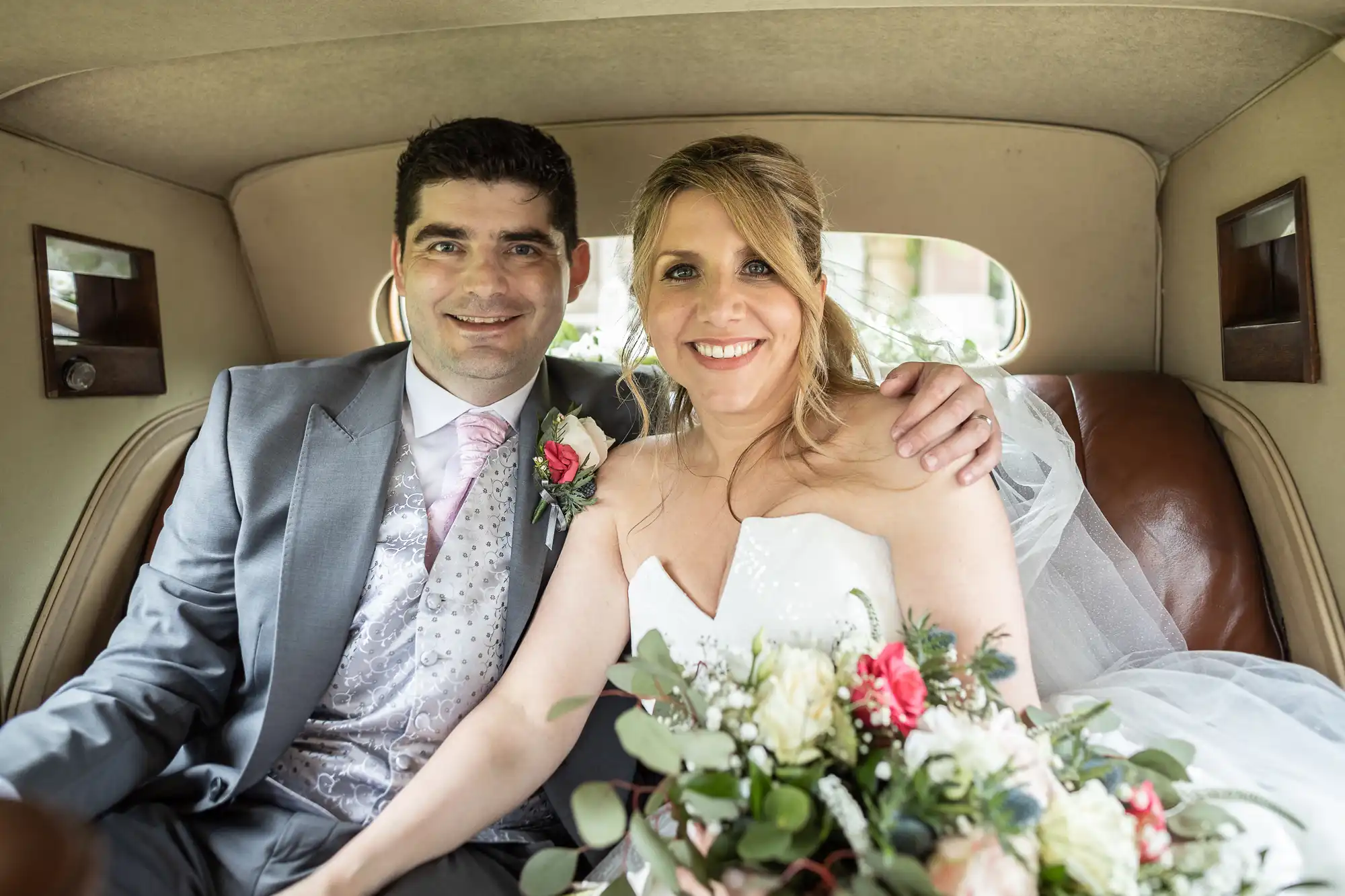 A bride and groom dressed in formal wedding attire are sitting together in the back seat of a vintage car, smiling at the camera while holding a bouquet of flowers.