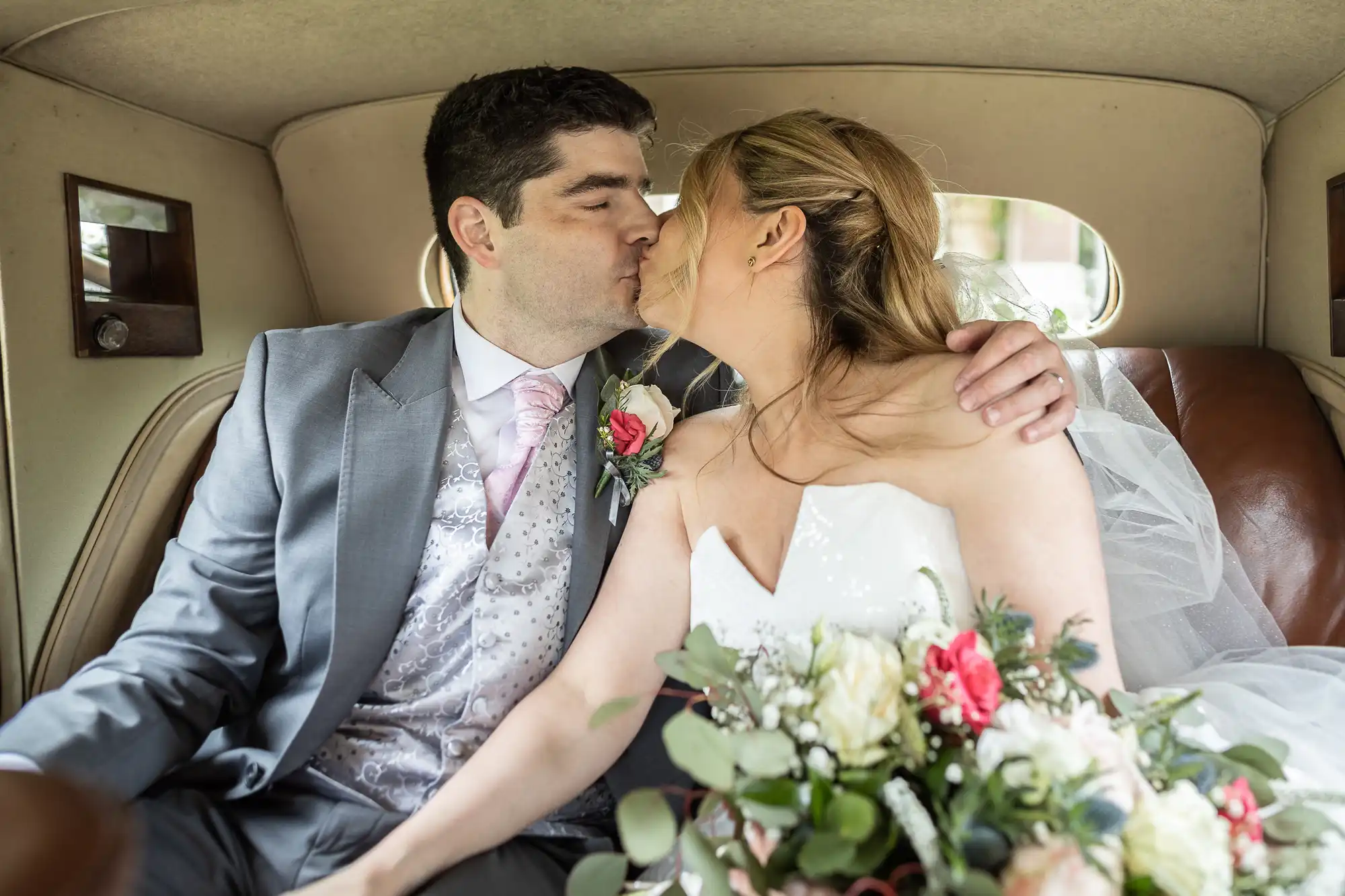 A bride and groom wearing formal wedding attire share a kiss while sitting in the backseat of a car, with the bride holding a bouquet of flowers.