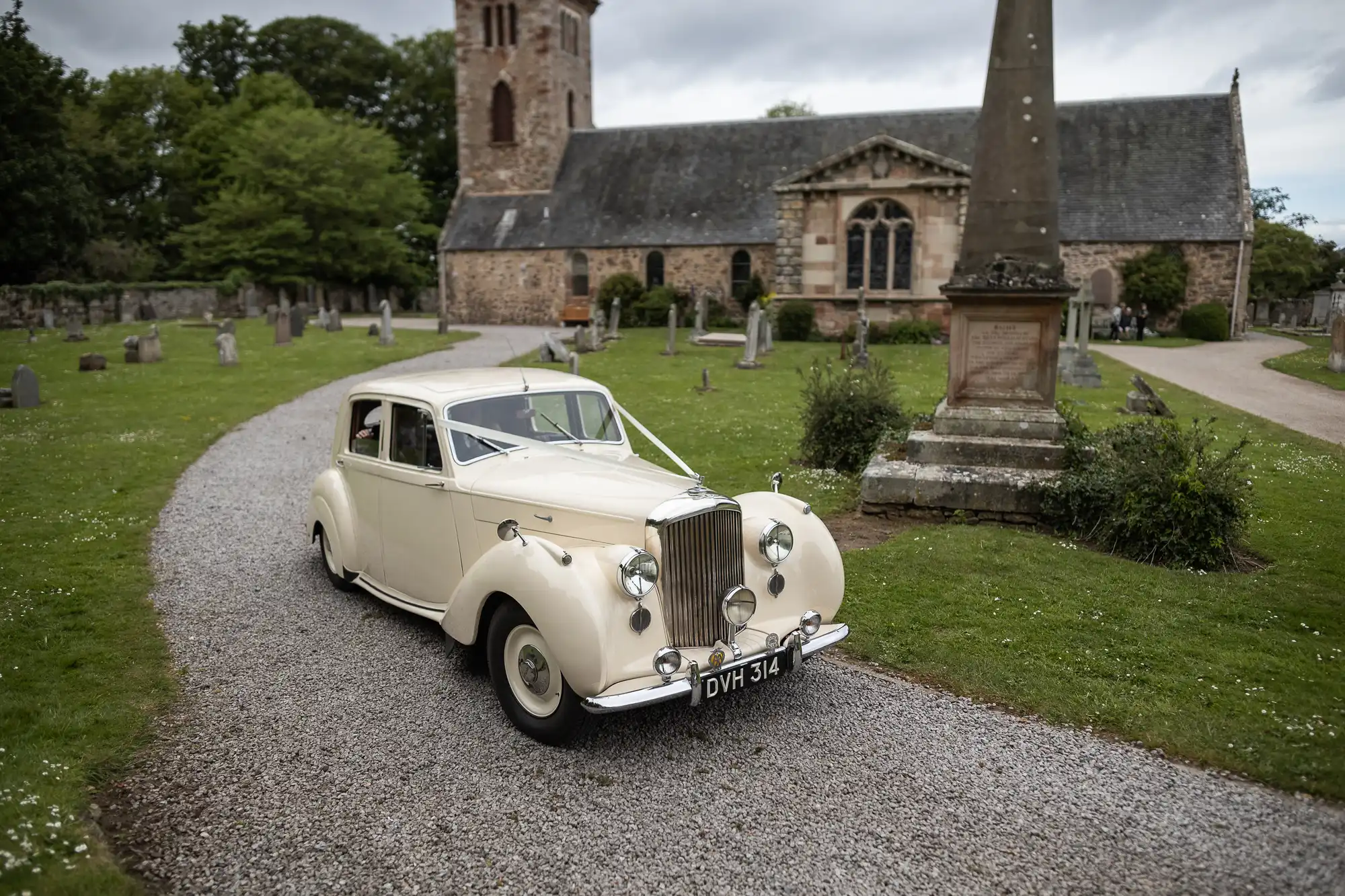 A vintage cream-colored car with the license plate "DWM 314" is parked on a gravel path in front of a historic stone church and cemetery.