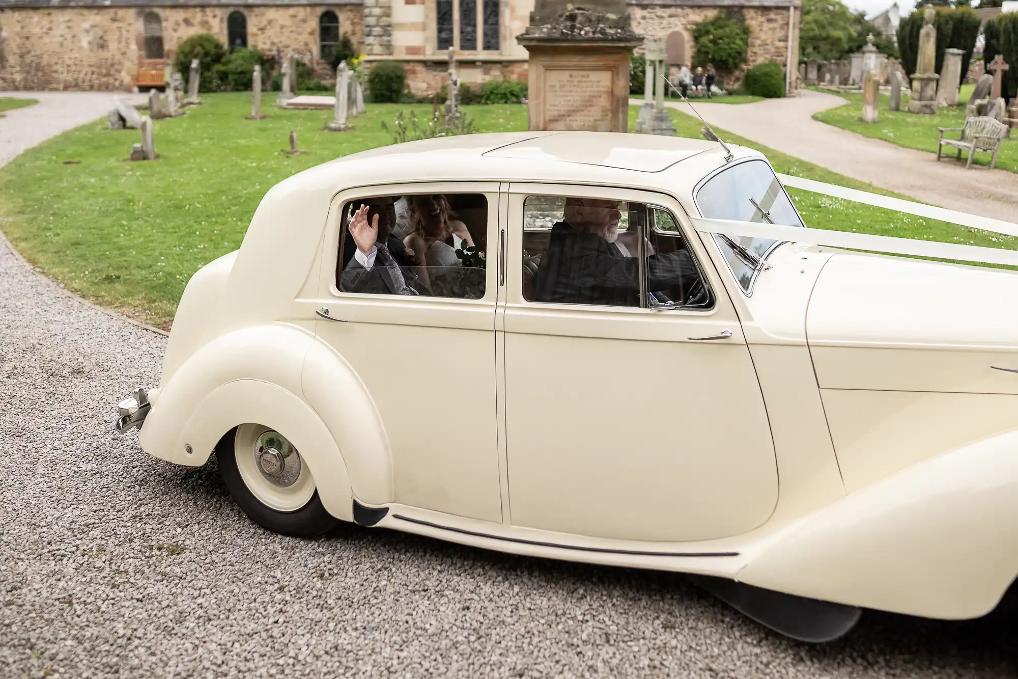 A classic white car with passengers inside drives through a cemetery. A person seated in the back seat waves.