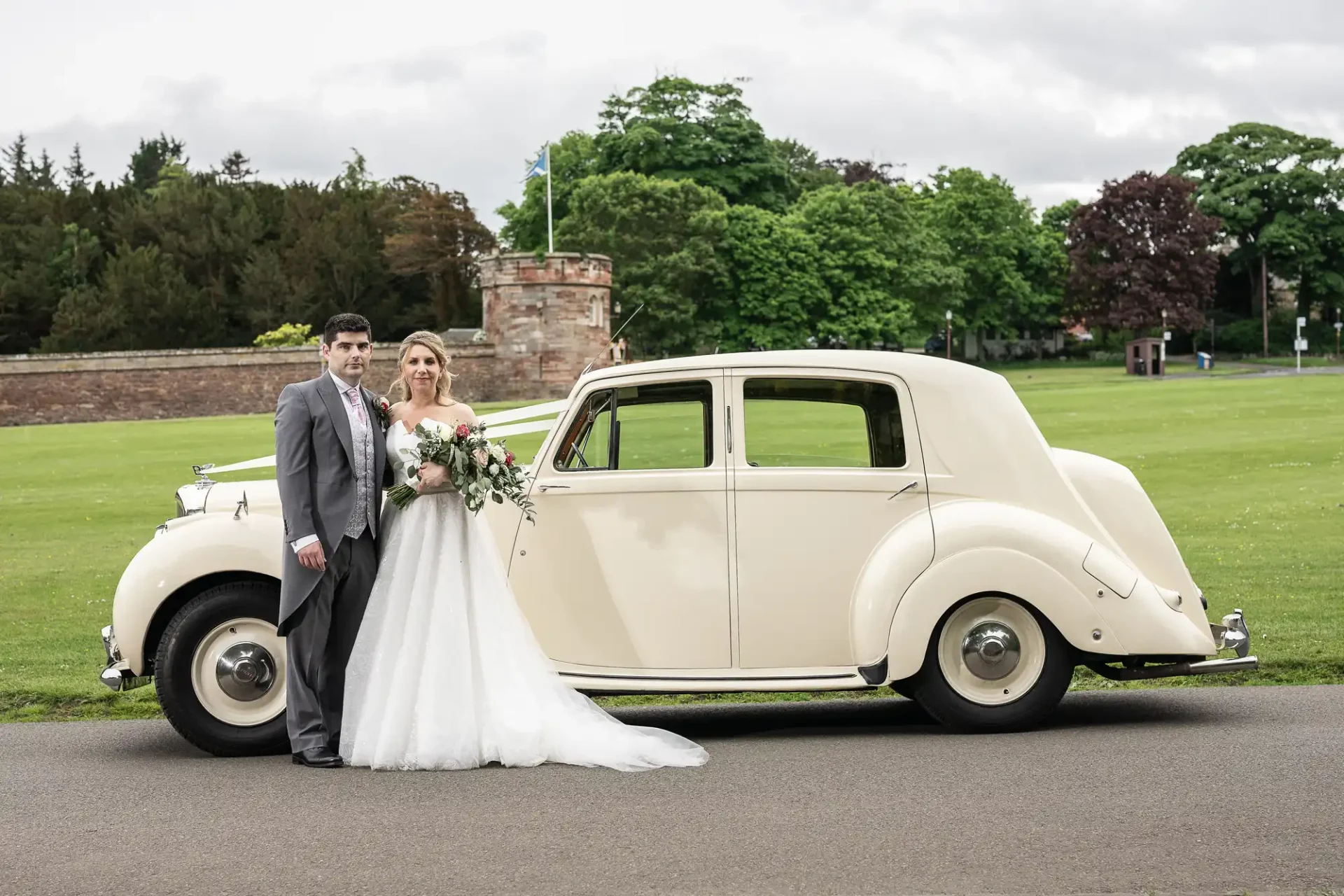 Dirleton Kirk wedding photographer: A bride and groom stand next to a vintage cream-colored car, posing for a photo in front of a grassy area with trees and a stone structure in the background.