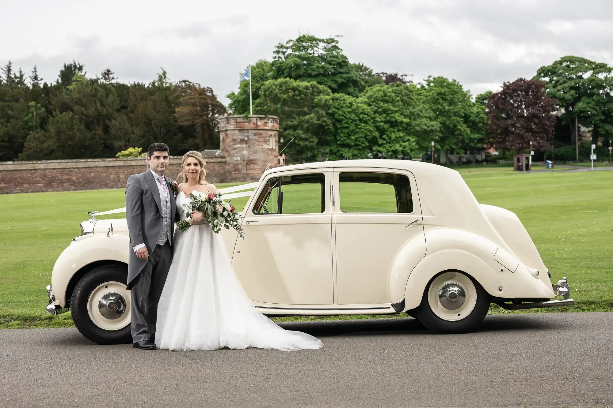 A bride and groom stand next to a vintage cream-colored car, posing for a photo in front of a grassy area with trees and a stone structure in the background.