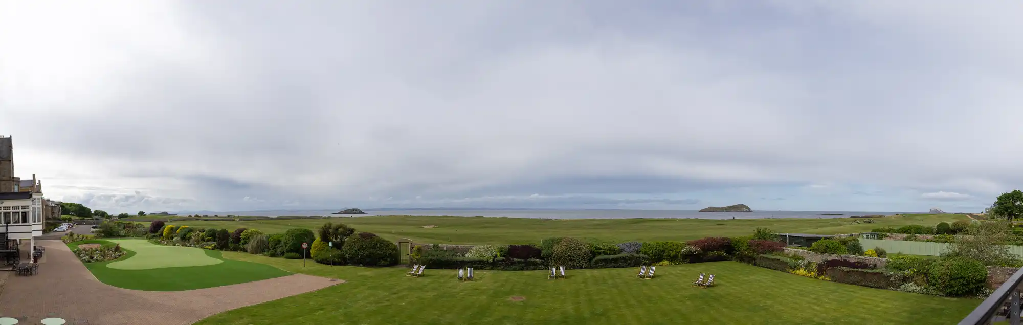 A spacious, grassy landscape with lounge chairs situated near a small putting green. The scene overlooks a vast, flat field with two small hills in the distance under a cloudy sky.