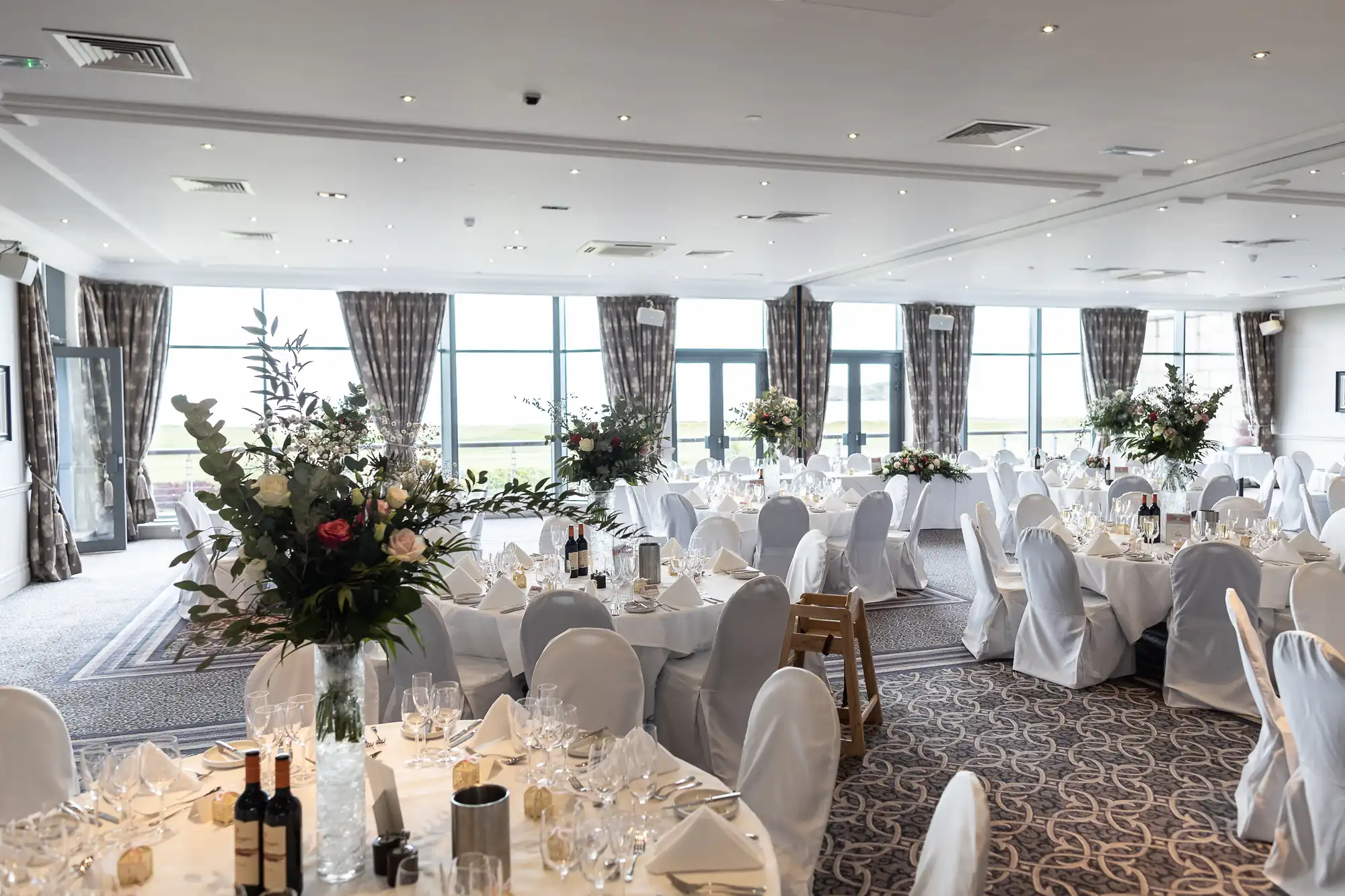 A banquet hall set up for an event with round tables draped in white tablecloths, surrounded by white chairs, and adorned with floral centerpieces and wine bottles.