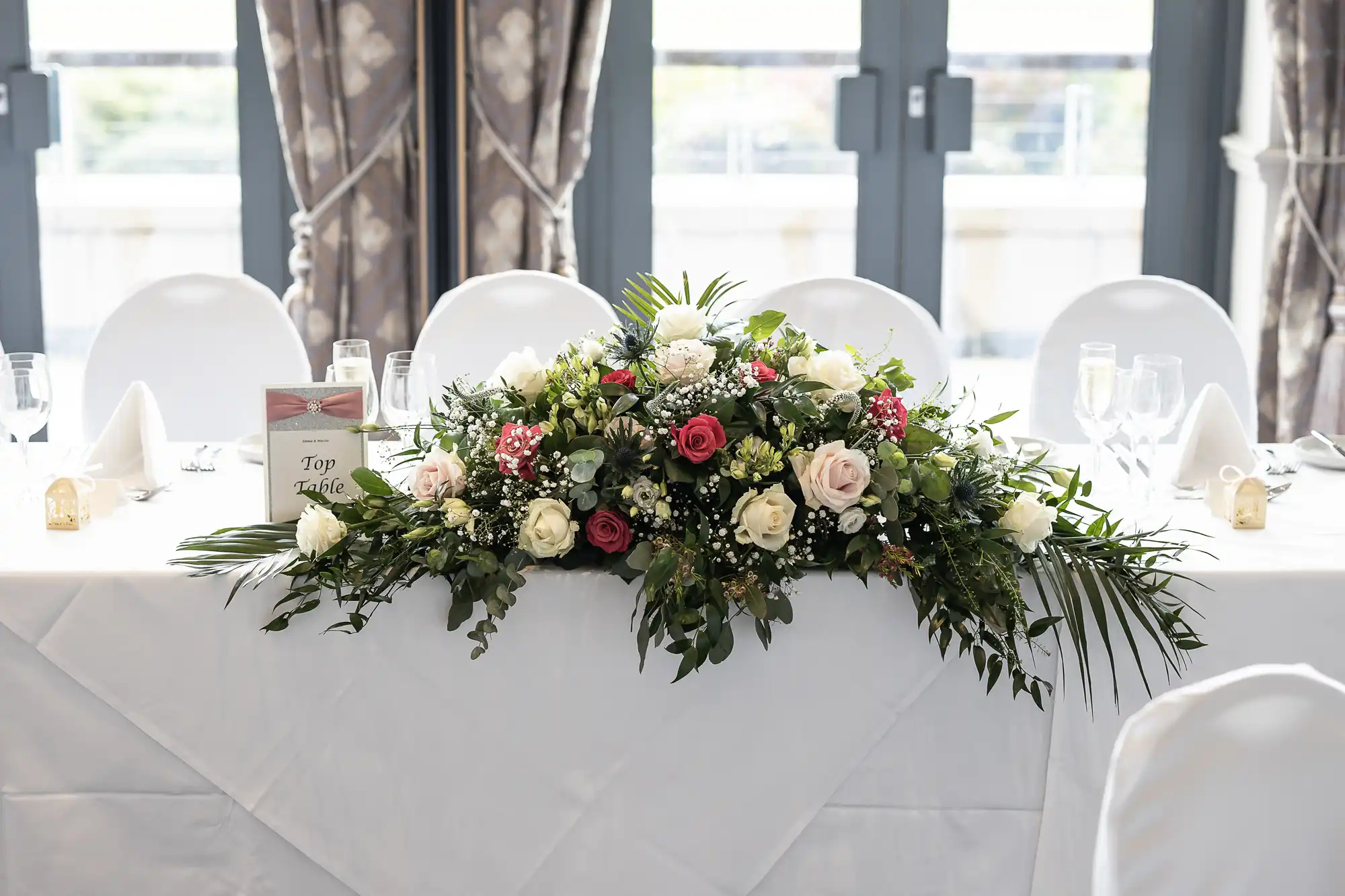 A table adorned with a floral centerpiece of white and pink roses, greenery, and white cloth napkins neatly folded on plates at each seating, set against a background of large windows with curtains.