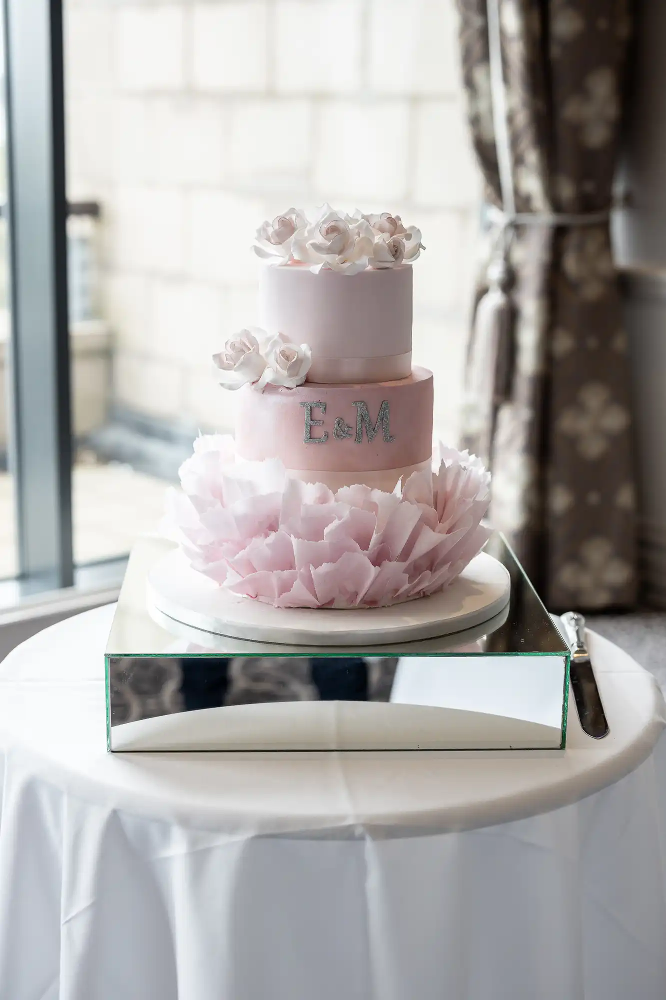 A two-tier pink wedding cake with white floral decorations and the initials "E & M" on a mirrored stand, placed on a round table.