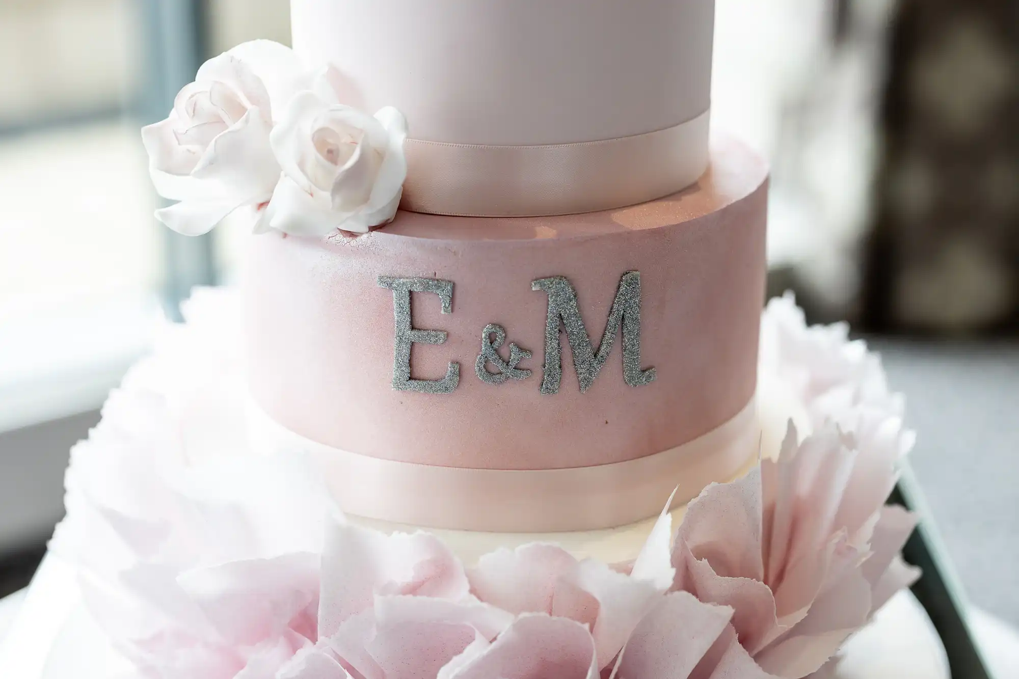 A white and pink tiered cake with the initials "E & M" in silver glitter on the middle tier, decorated with white flowers and pink petal-like embellishments at the base.