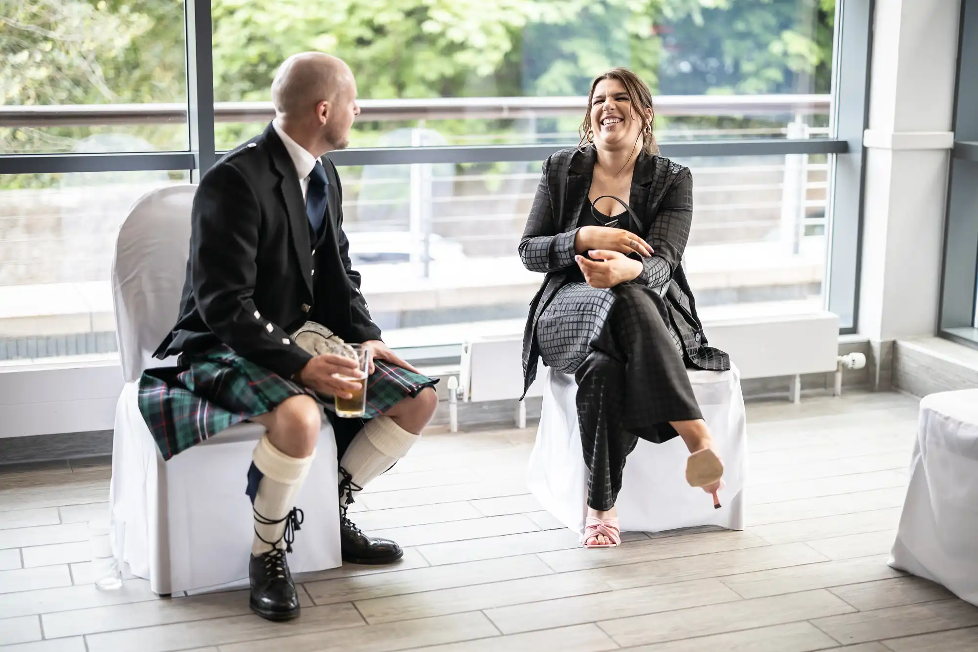 A man in a kilt and a woman in a plaid outfit sit on white chairs indoors, engaging in an animated conversation. The man holds a glass, and the background shows large windows with a view of trees.