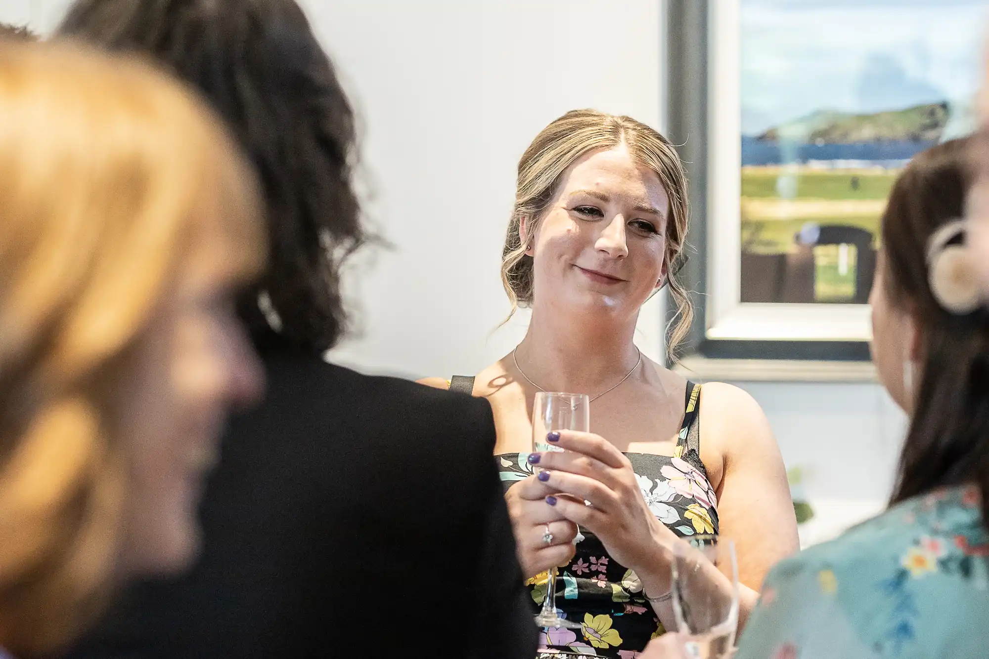 A woman in a floral dress holds a glass and smiles while conversing with others in a room decorated with framed artwork.