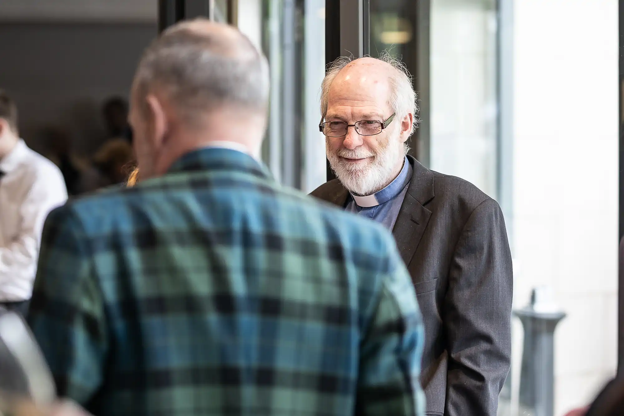 Two older men are conversing, with one facing towards the camera and the other wearing a plaid jacket and facing away. They appear to be in a well-lit indoor setting.