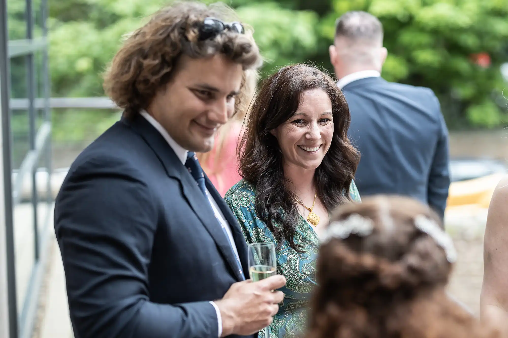 Two people are smiling and conversing at an outdoor event, with glasses in hand. They are dressed formally. Other attendees are visible in the background.