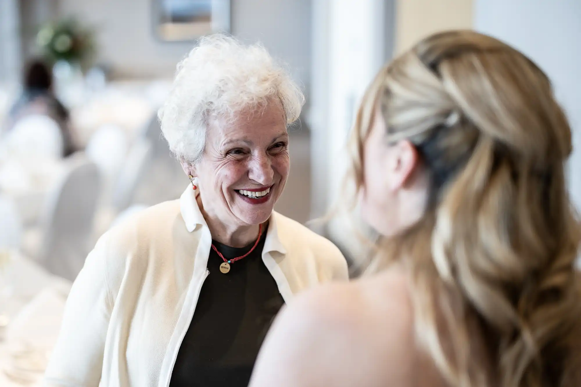 An elderly woman with short white hair smiles at a younger woman with long blonde hair in an indoor setting.