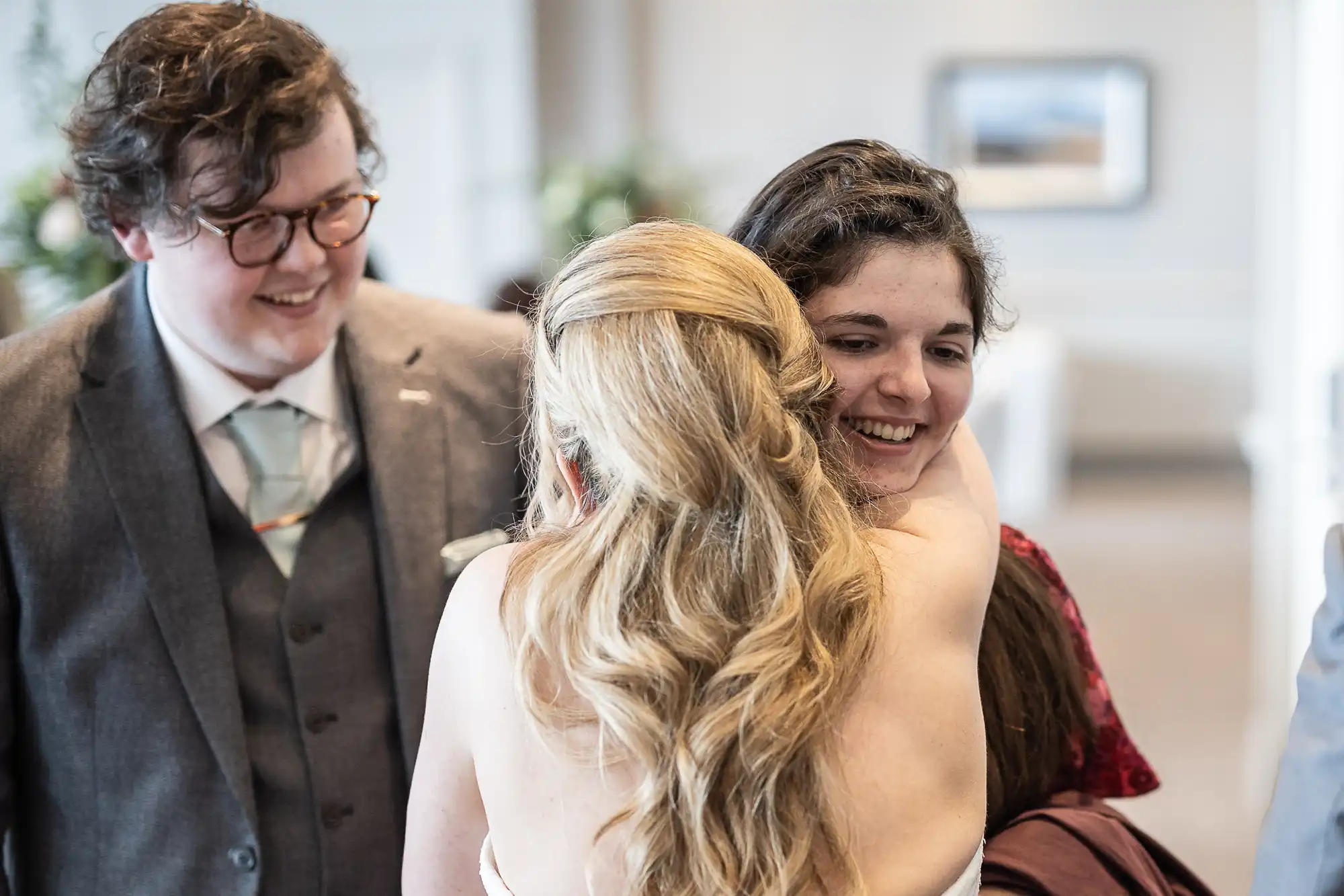 Three people interacting; a person in a white dress with blonde hair hugs a person with long dark hair and a red dress. A person in a gray suit with glasses stands by, smiling.