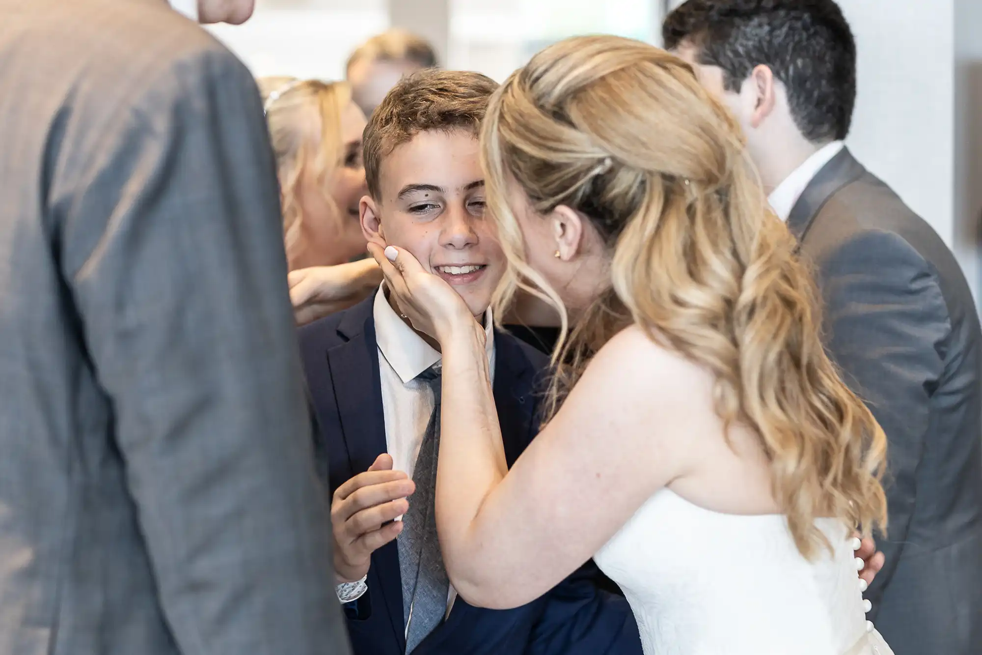 Woman in a white dress affectionately pinching the cheek of a young boy in a suit, surrounded by other formally dressed people.