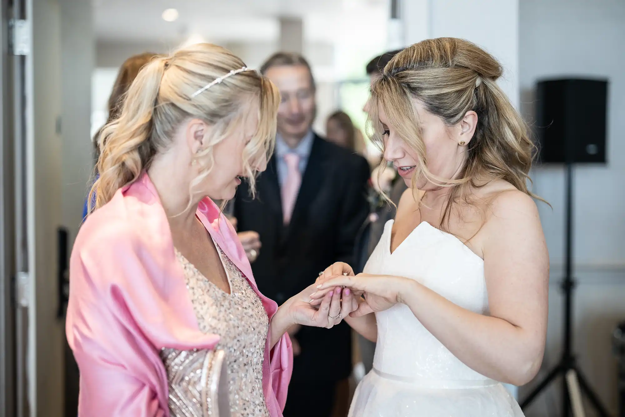 Two women are standing together at an event; one in a white dress and the other in a pink dress. Both appear to be examining a ring on the woman in the white dress. Other blurred figures are seen in the background.