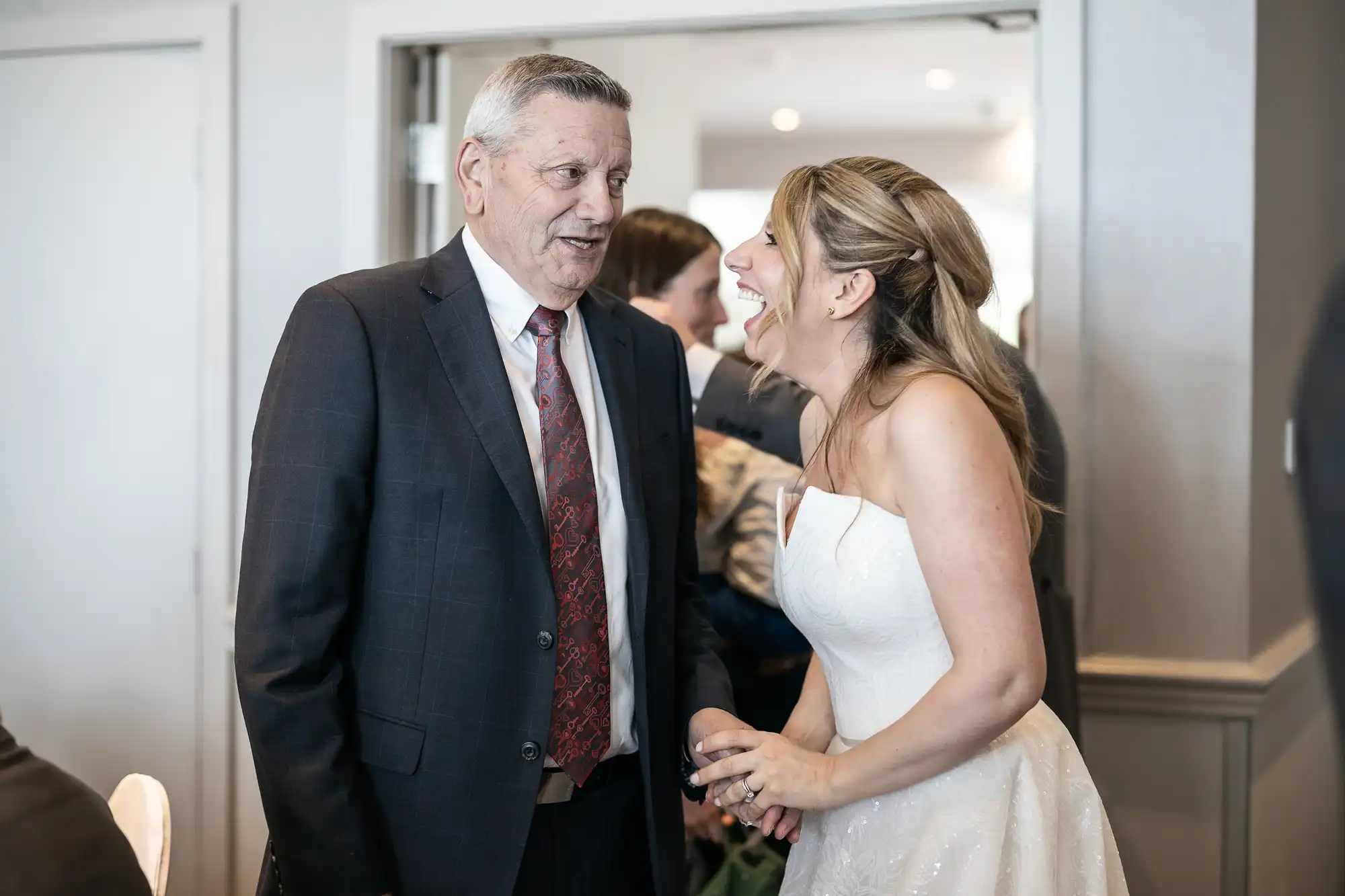 A man in a suit and tie converses with a woman in a white dress, who is smiling and holding a glass, in a well-lit room with people in the background.