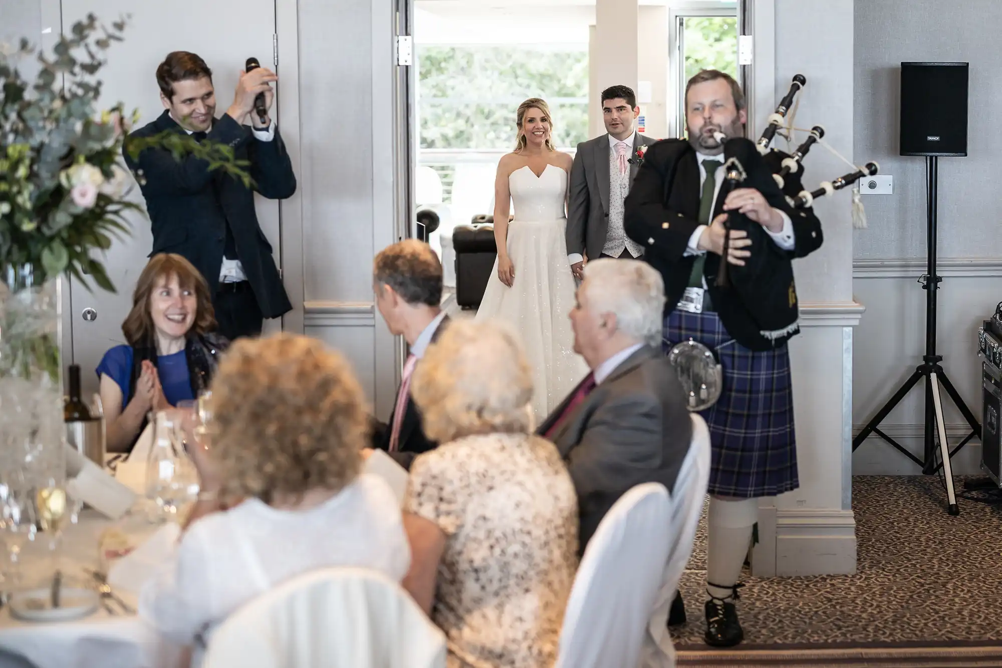 A wedding scene: a couple is entering a room with the bride in a white dress, followed by a man in a suit. A bagpiper in a kilt is playing, while seated guests look on and a man takes a photo.