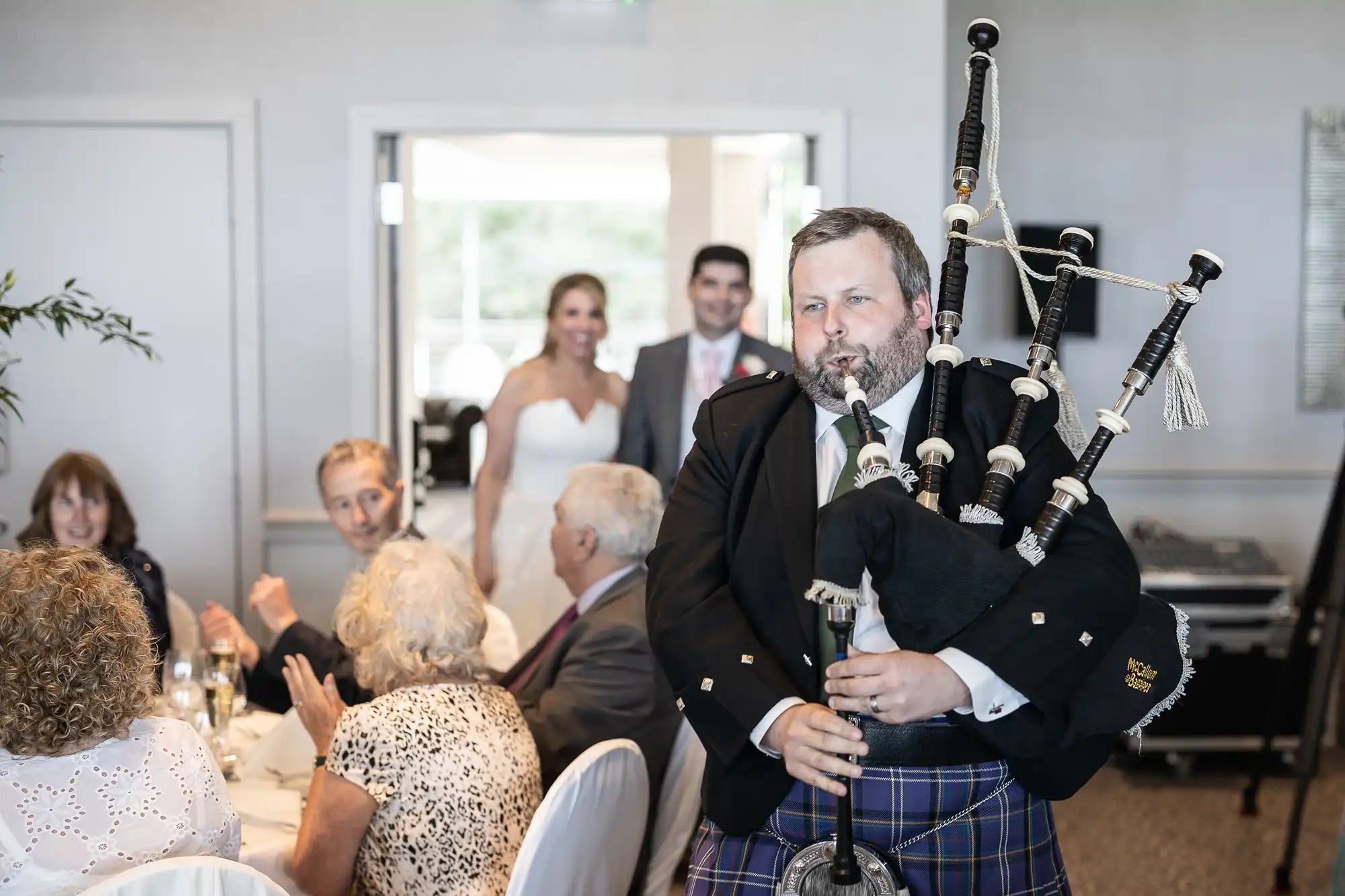 A man in traditional Scottish attire plays the bagpipes at an indoor event. Guests are seated at round tables, and a couple, presumably newlyweds, smile in the background.